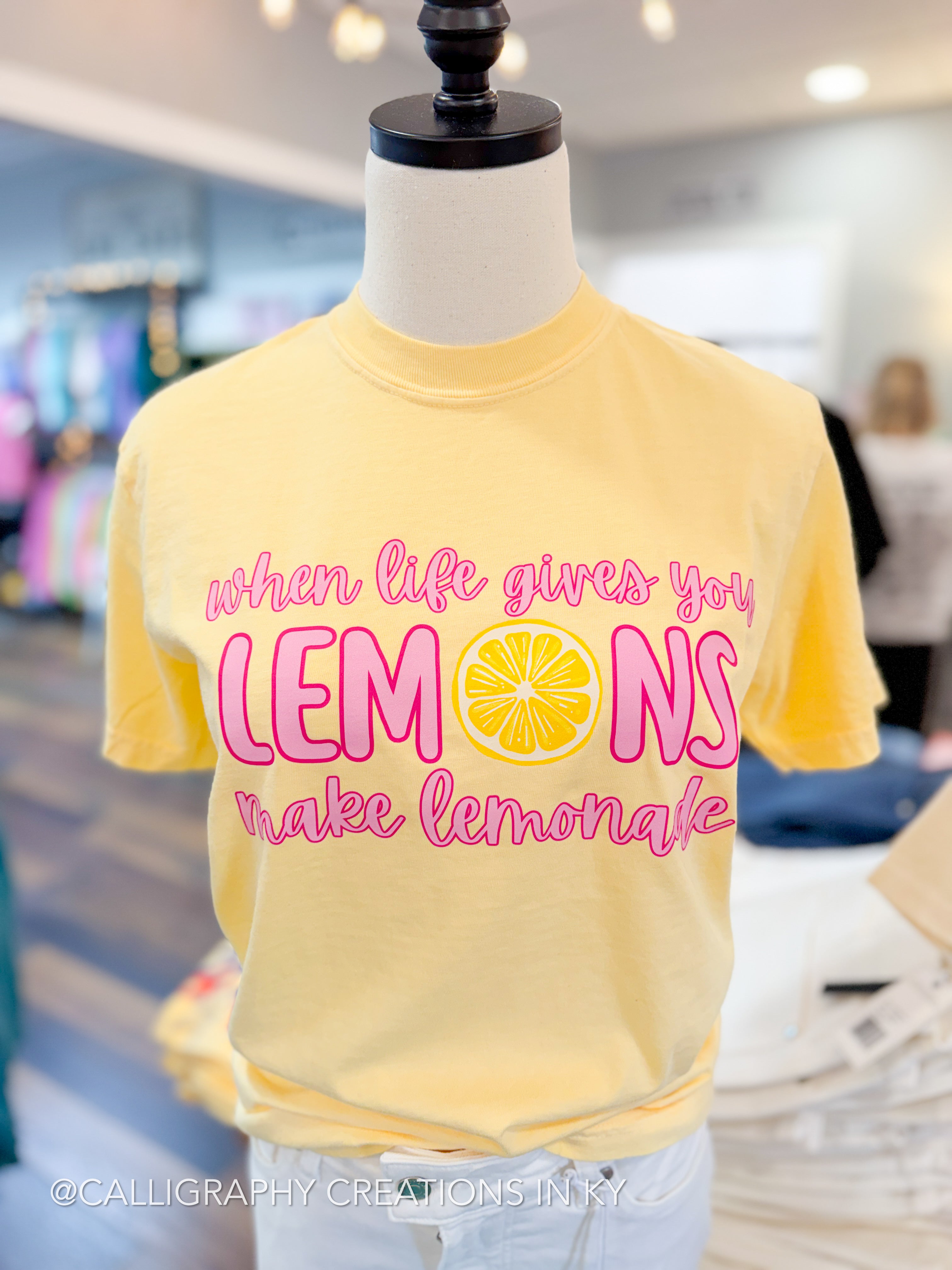 Yellow t-shirt with 'When life gives you lemons make lemonade' text on a mannequin in a store setting.