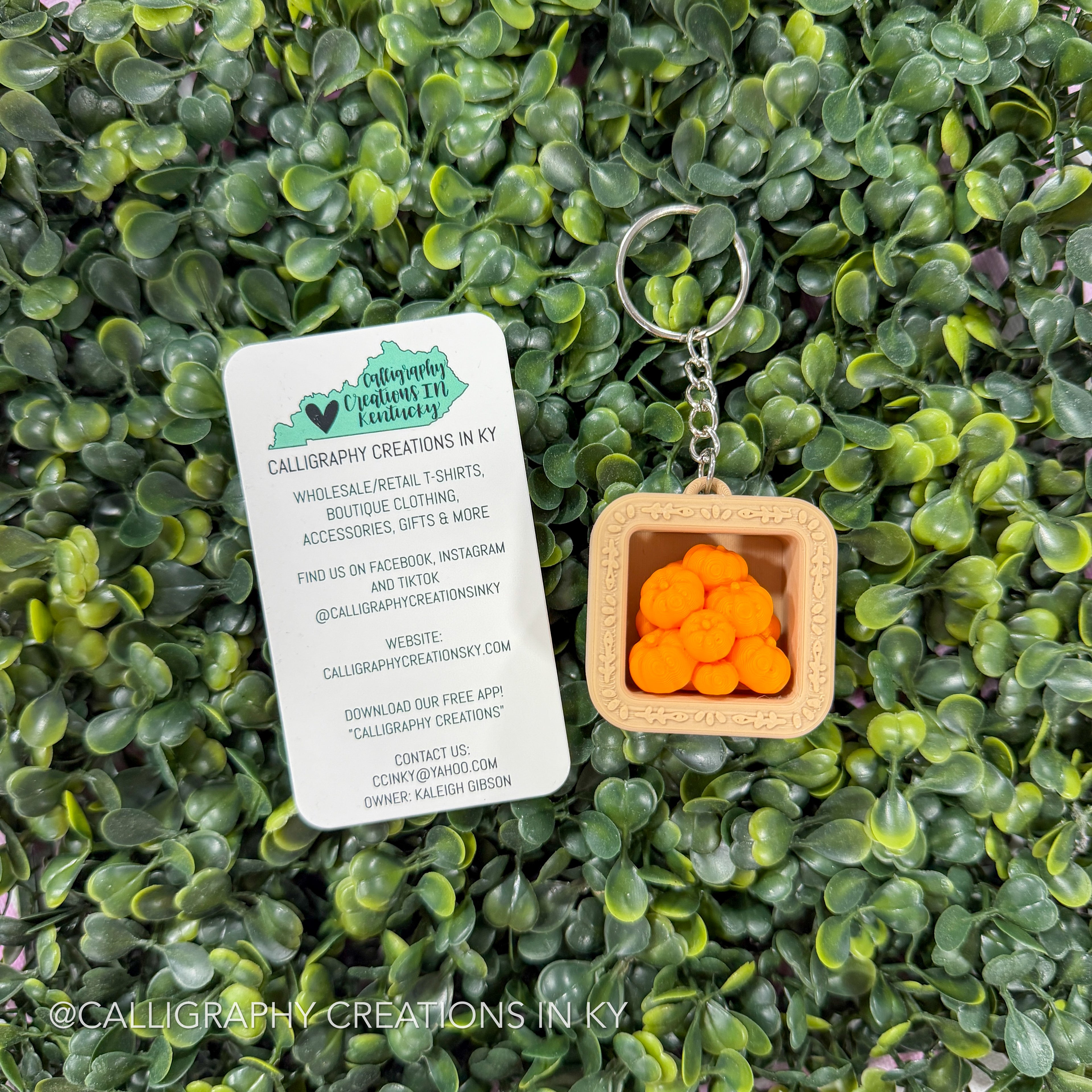 A square wooden calligraphy keychain with a silver metal ring and chain, featuring a light wood tray filled with small orange calligraphy characters, displayed on a background of green leaves next to a white brand card.