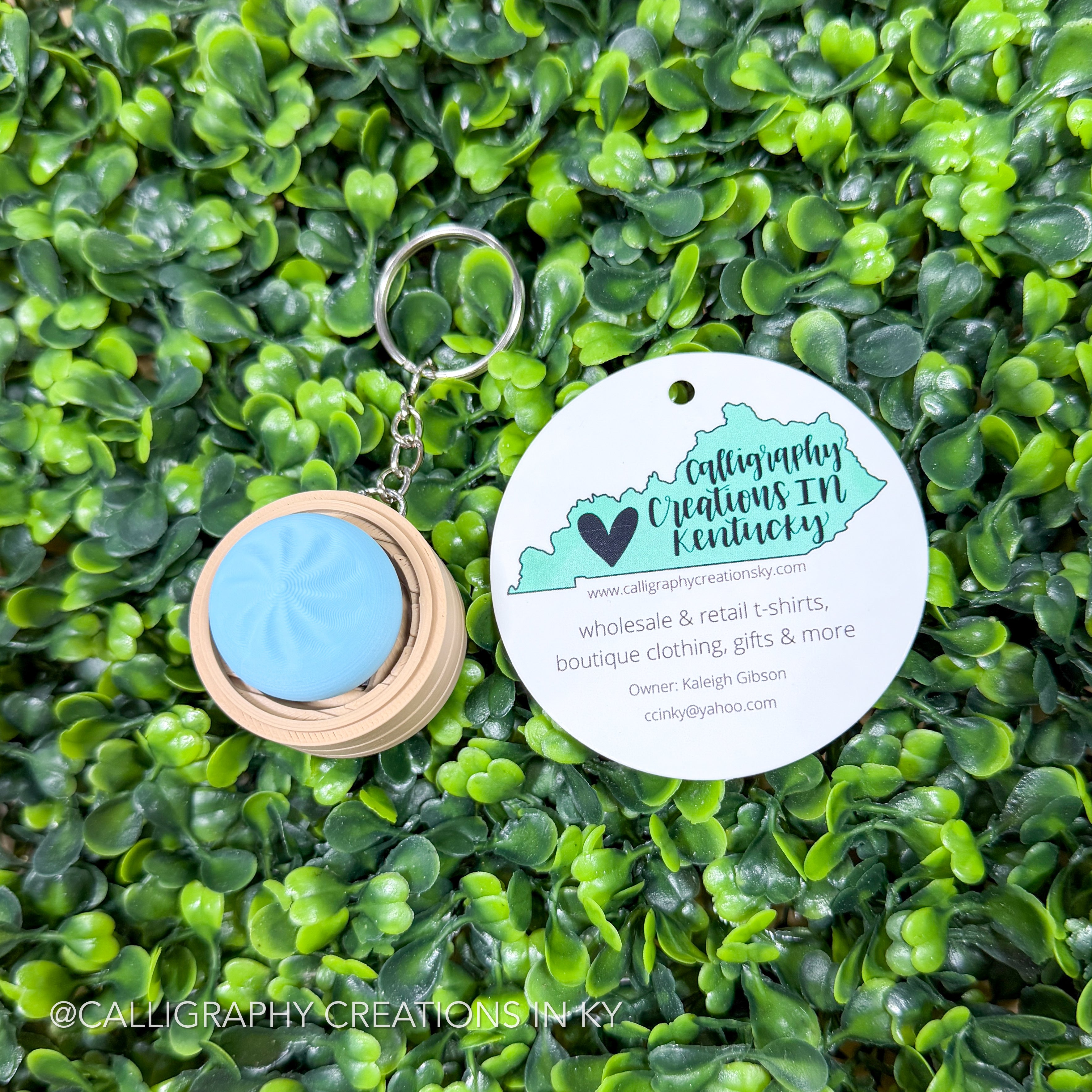 Keychain with a blue dumpling fidget clicker and wooden border on a green leafy background, featuring Calligraphy Creations in Kentucky branding.