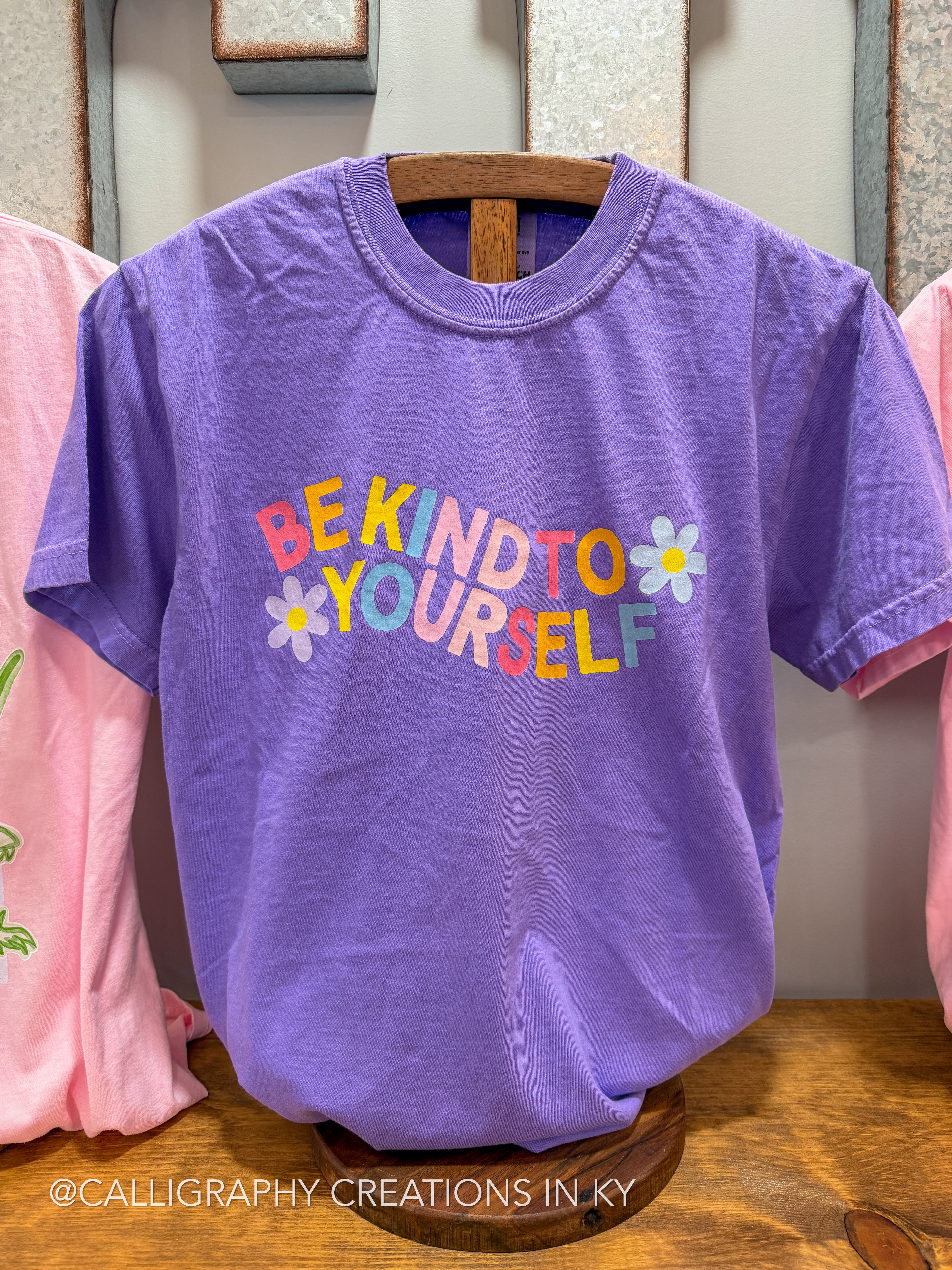 Purple t-shirt with 'Be Kind to Yourself' text on a wooden hanger.