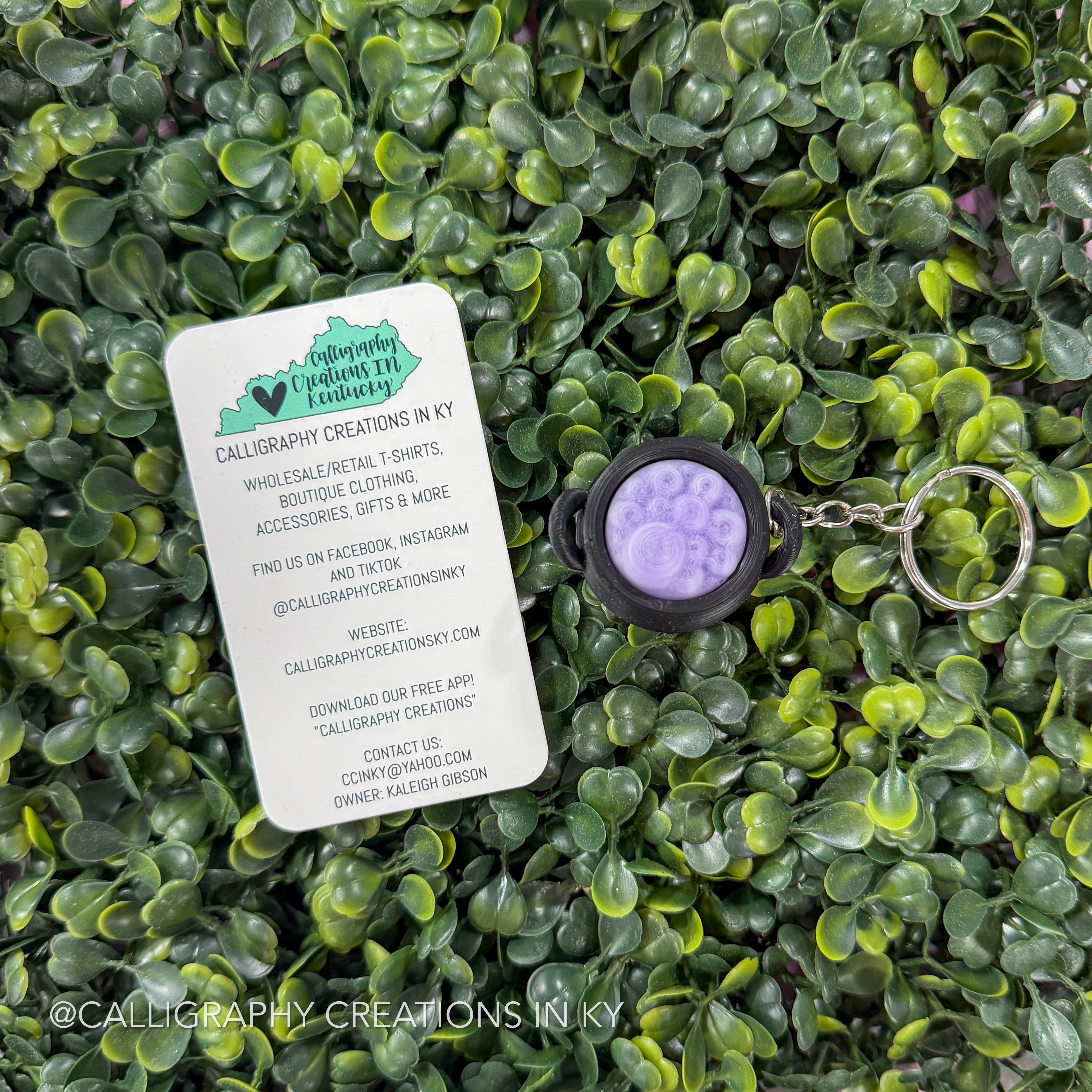 A calligraphy-themed keychain with a round purple and lavender design in a black bezel, shown next to a white brand card on a background of green artificial leaves.