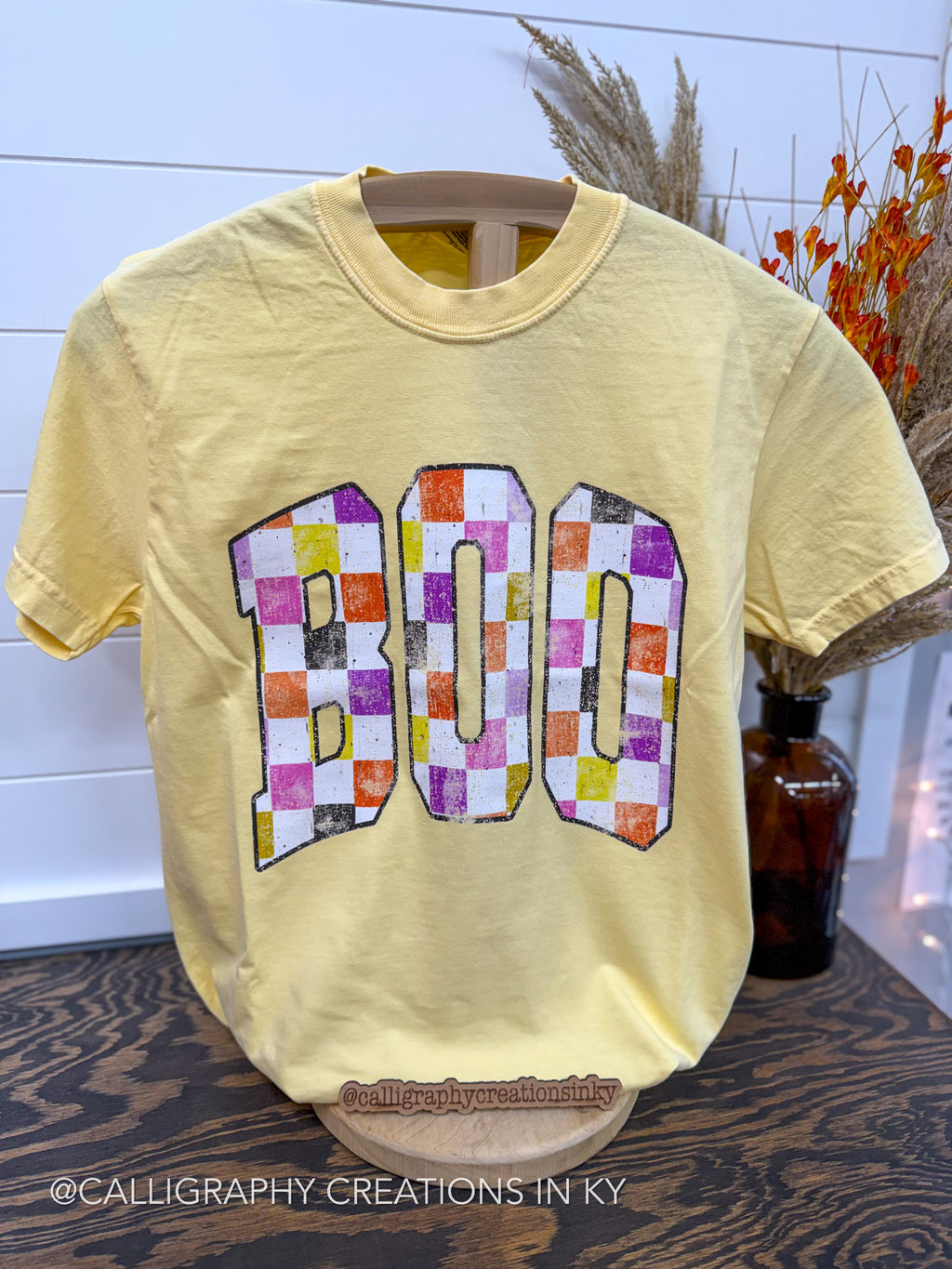 A pale yellow short-sleeve T-shirt displayed on a wooden hanger, featuring a large 'BOO' graphic across the chest in a multicolor checkerboard pattern. The shirt is photographed indoors on a dark wood surface with a white shiplap wall and dried floral decor in the background.