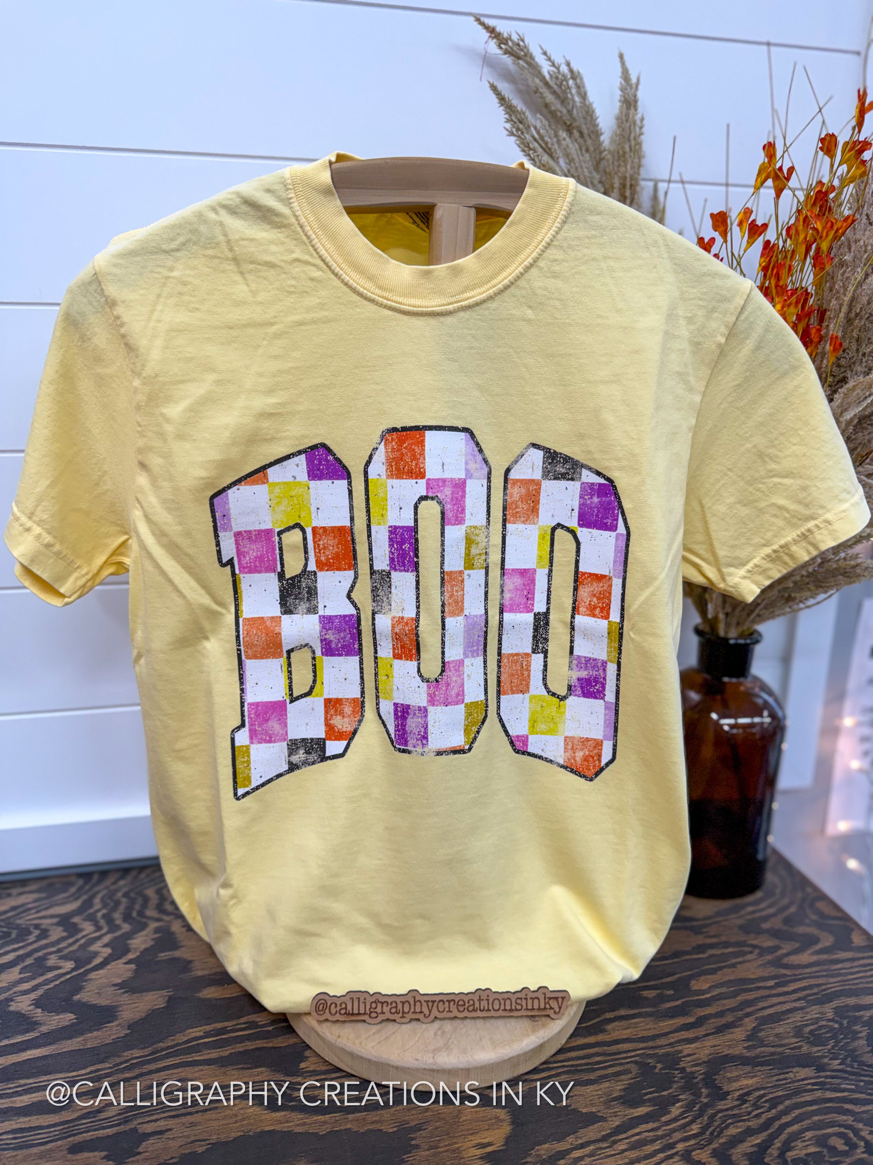 A pale yellow short-sleeve T-shirt displayed on a wooden hanger, featuring a large 'BOO' graphic across the chest in a multicolor checkerboard pattern. The shirt is photographed indoors on a dark wood surface with a white shiplap wall and dried floral decor in the background.