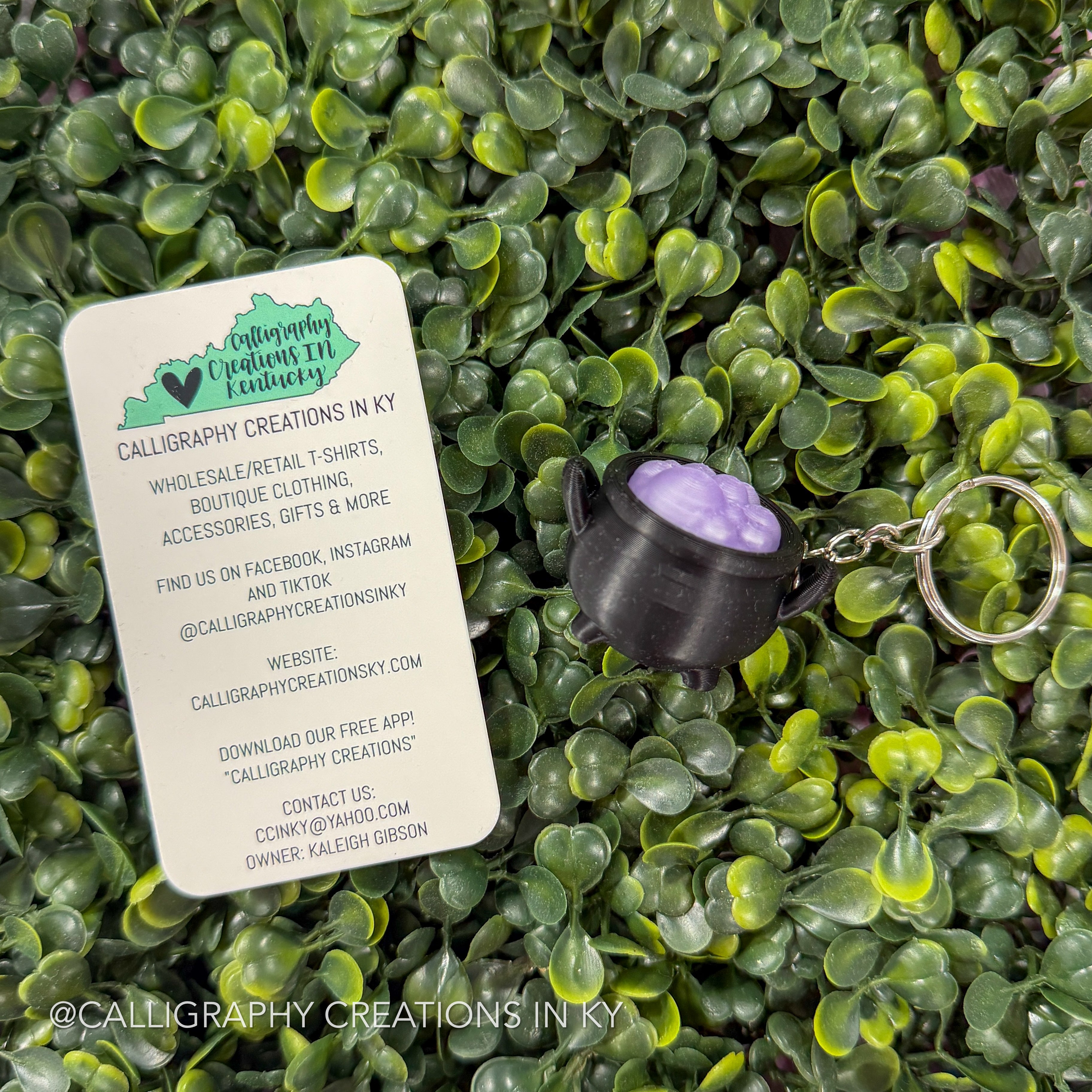 A small black cauldron-shaped keychain with a purple lid sits on green leafy foliage next to a white branded card that reads 'Calligraphy Creations in KY' and lists wholesale/retail items and social media details.