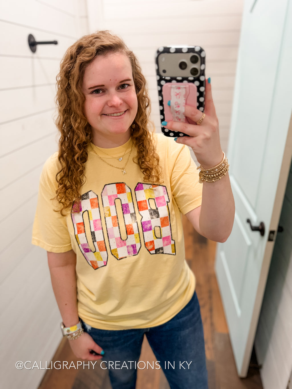 A woman taking a mirror selfie in a bathroom wearing a yellow t-shirt with a colorful checkered 'BOO' graphic. The photo is taken in a bright bathroom with white shiplap walls and wood floors.