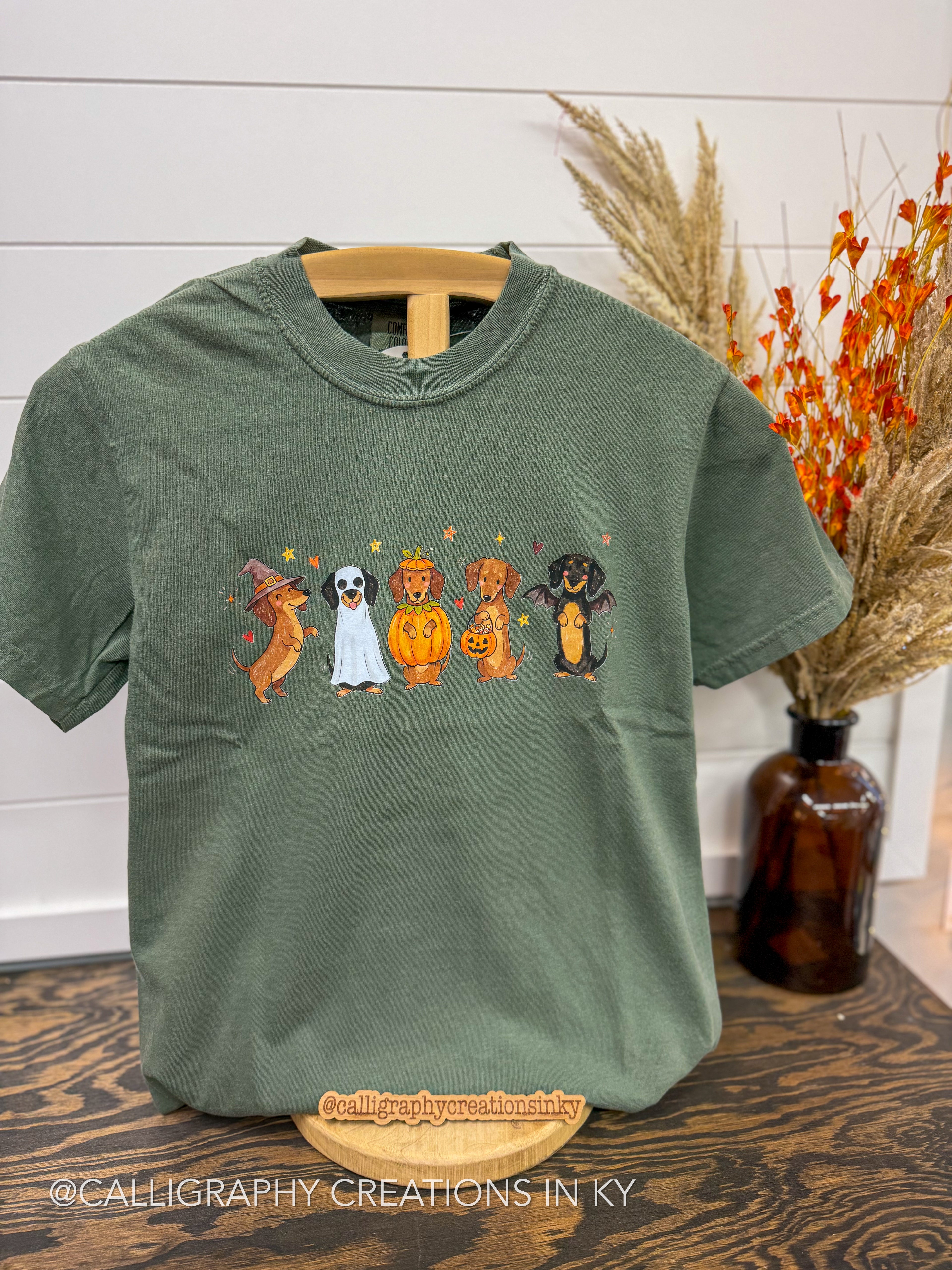 An olive green short-sleeve t-shirt on a wooden hanger, featuring a graphic of four dogs dressed in Halloween costumes. The background includes a white shiplap wall, a wooden table, and a decorative brown glass vase with dried orange leaves.