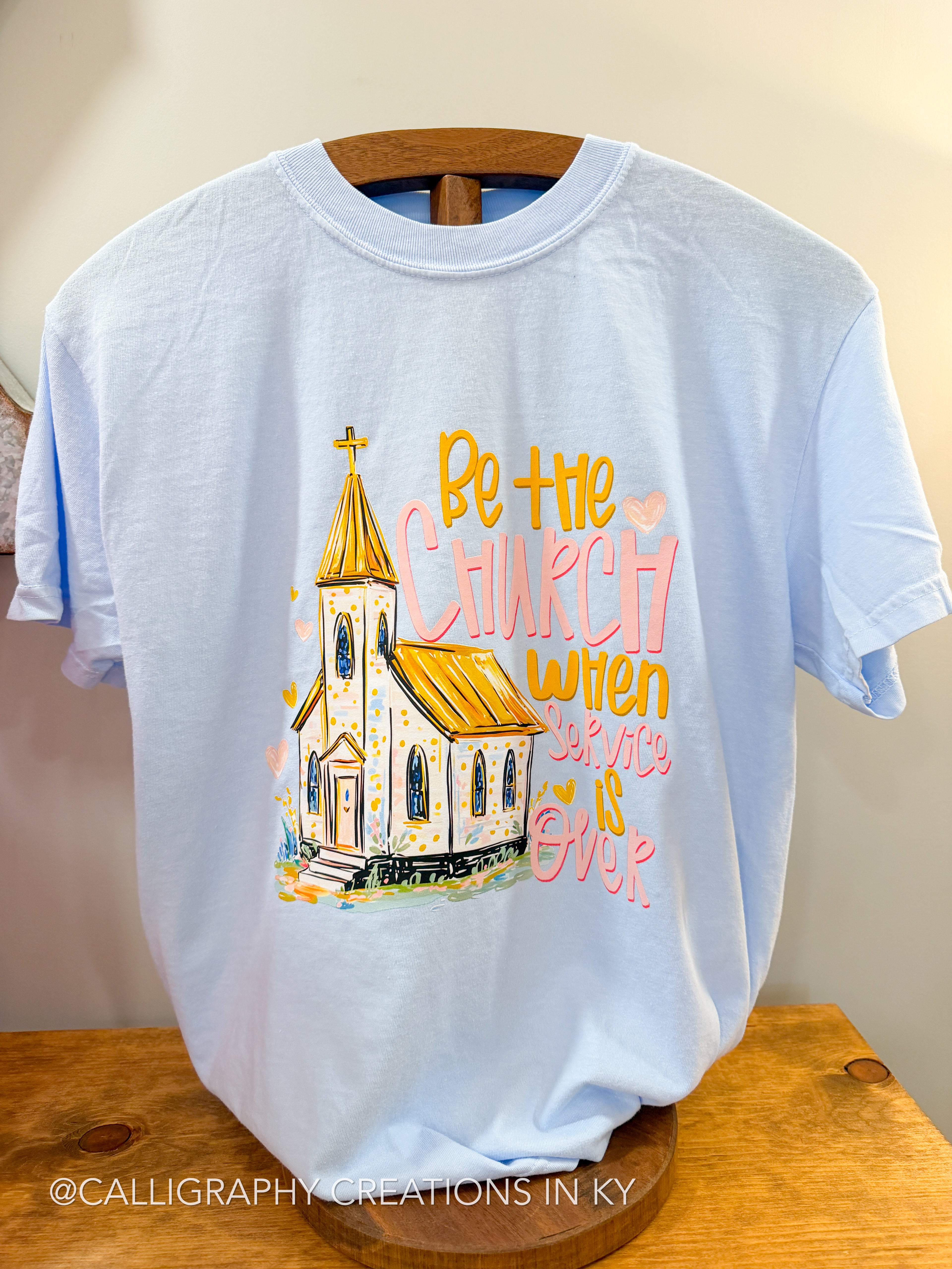 Light blue t-shirt with a church graphic and text on a wooden hanger against a beige wall.