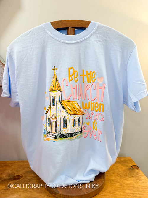 Light blue t-shirt with a church graphic and text on a wooden hanger against a beige wall.