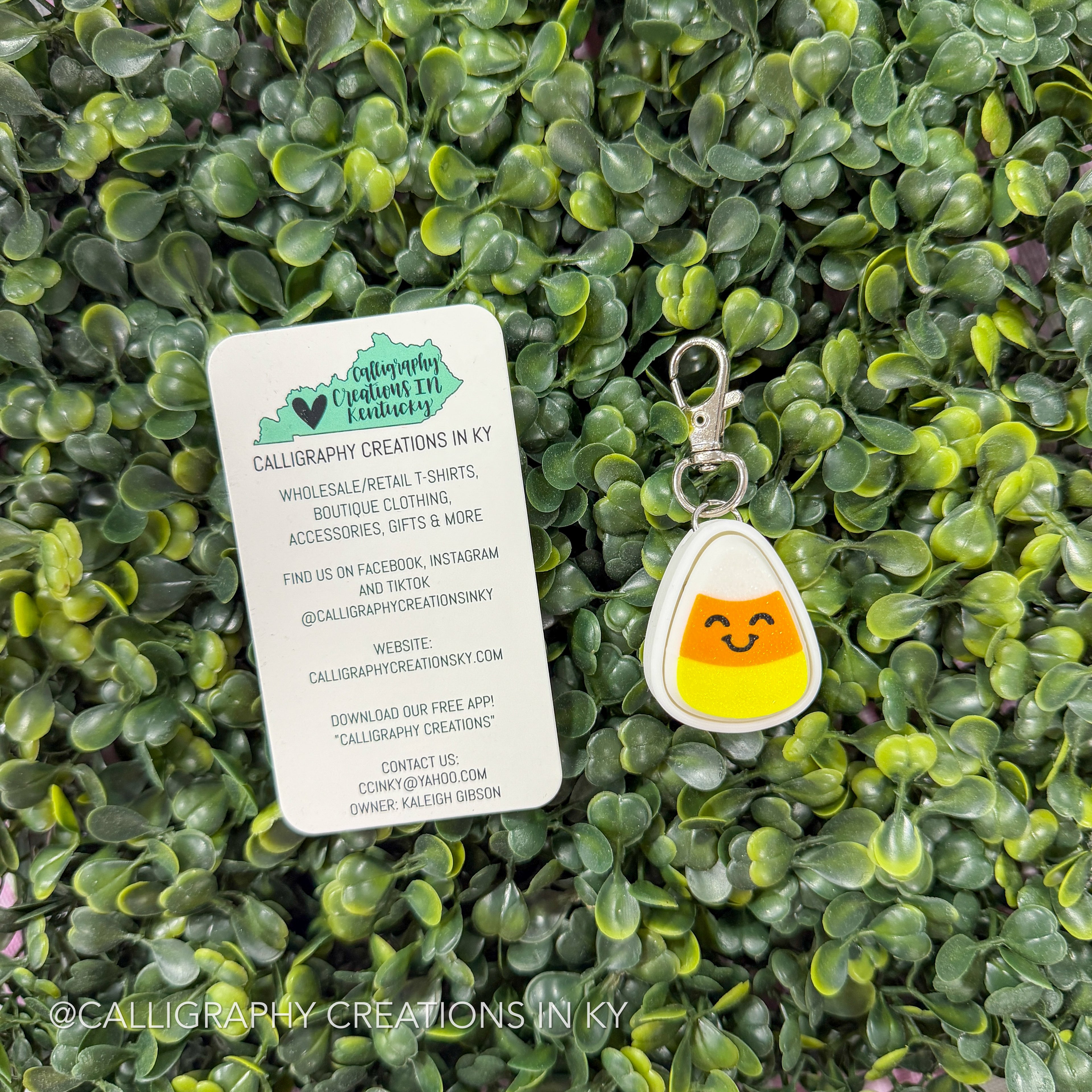 A small candy corn keychain charm with a smiling face is displayed on a white branded card, set against a background of green artificial foliage. The card shows the brand 'Calligraphy Creations in KY'.