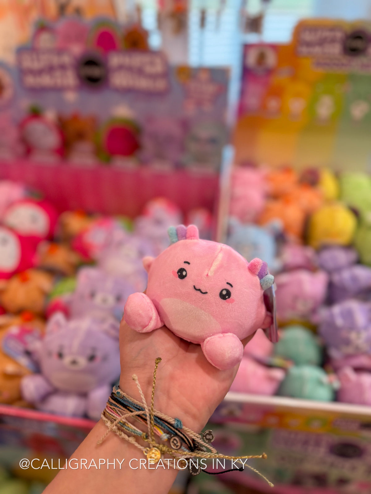 A hand holding a small pink plush toy with a cute face and horns, with a blurred arcade display of other plushies in the background.