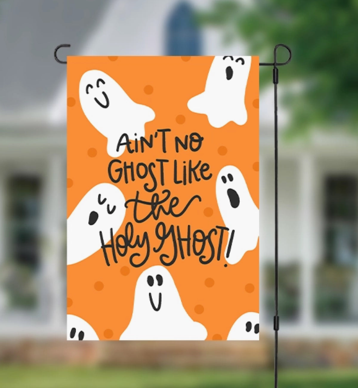An orange Halloween garden flag hanging from a black metal stand, featuring white cartoon ghosts and the text 'Ain't no ghost like the Holy Ghost!' against a blurred outdoor house background.
