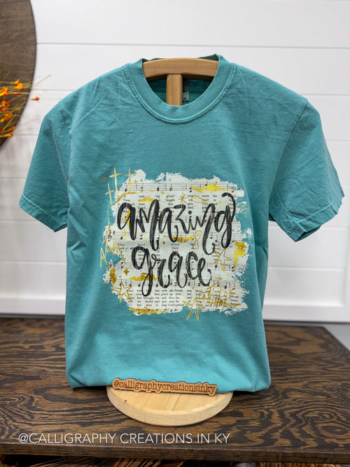 A teal short-sleeve graphic T-shirt hanging on a wooden hanger against a white paneled wall, with a large front print reading “amazing grace” in black script over a white and gold textured background. The shirt is displayed on a wooden stand on a dark wood table, with a watermark at the bottom.