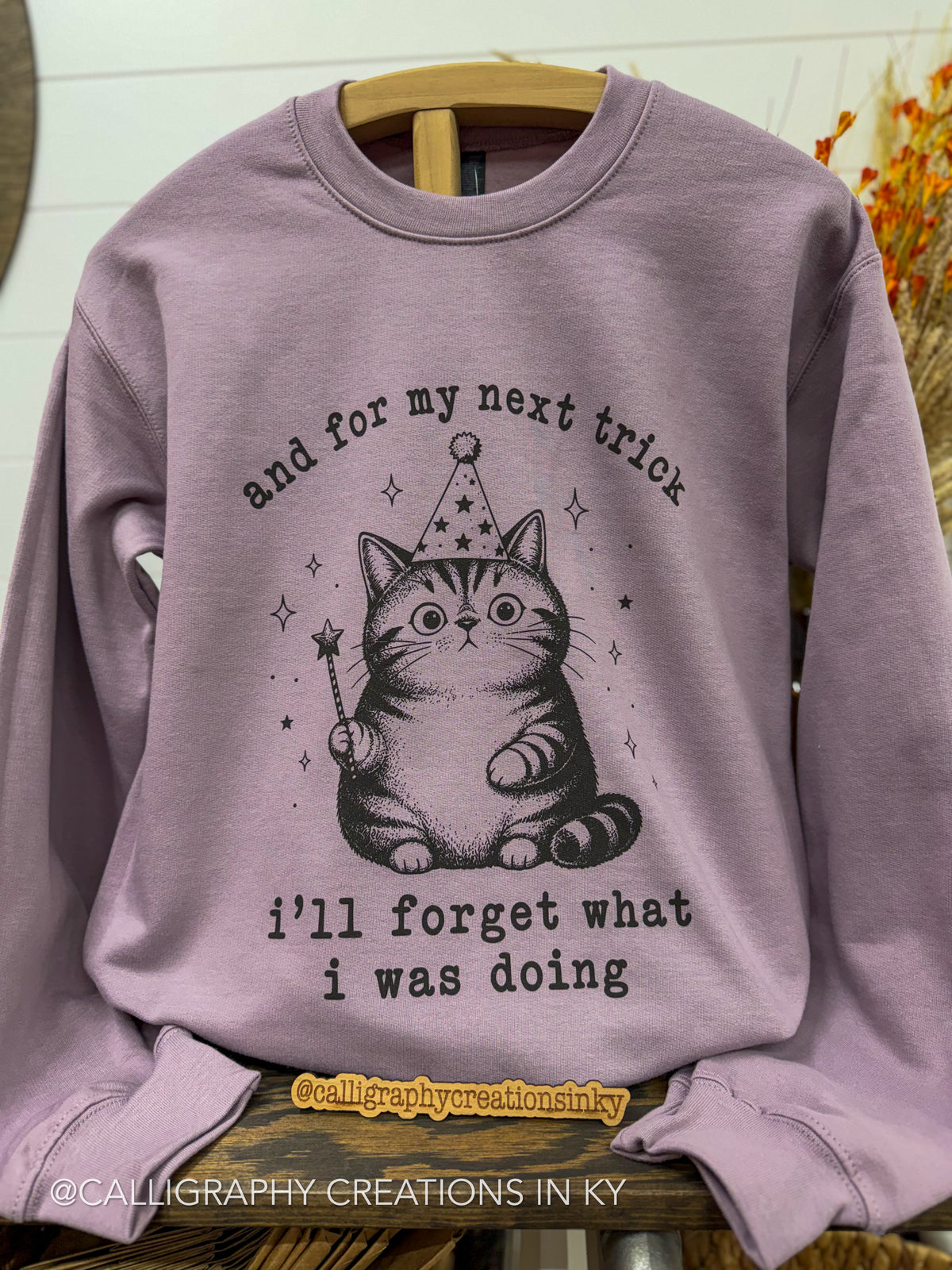 A lavender crewneck sweatshirt hanging on a wooden hanger, featuring a black graphic of a cat wearing a party hat and holding a wand, with the text 'and for my next trick i'll forget what i was doing'.