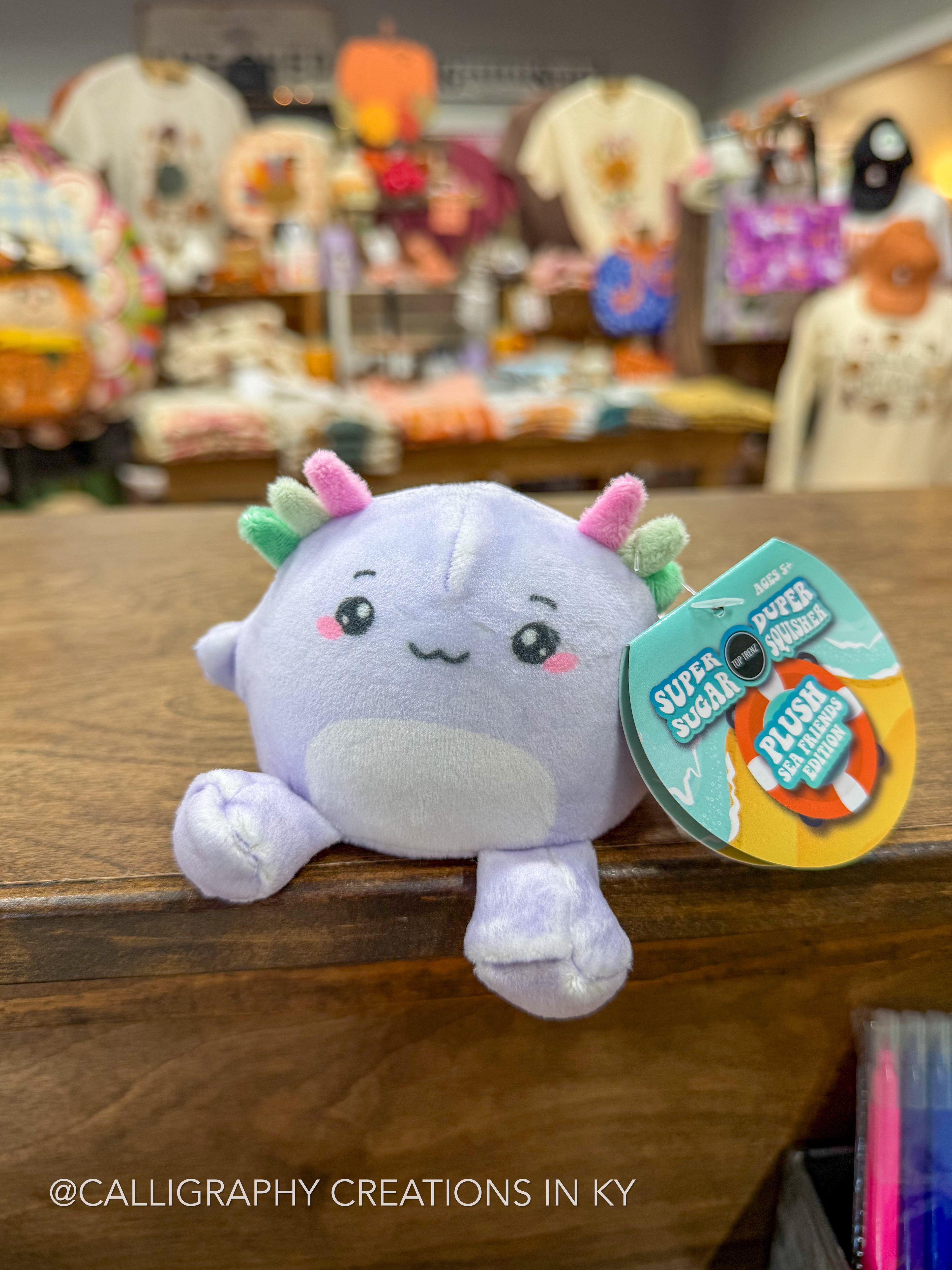 A small plush toy with a pale lavender body, pink and green accents, and a hangtag reading 'Super Squishy' photographed on a wooden counter in a retail store with blurred merchandise in the background.