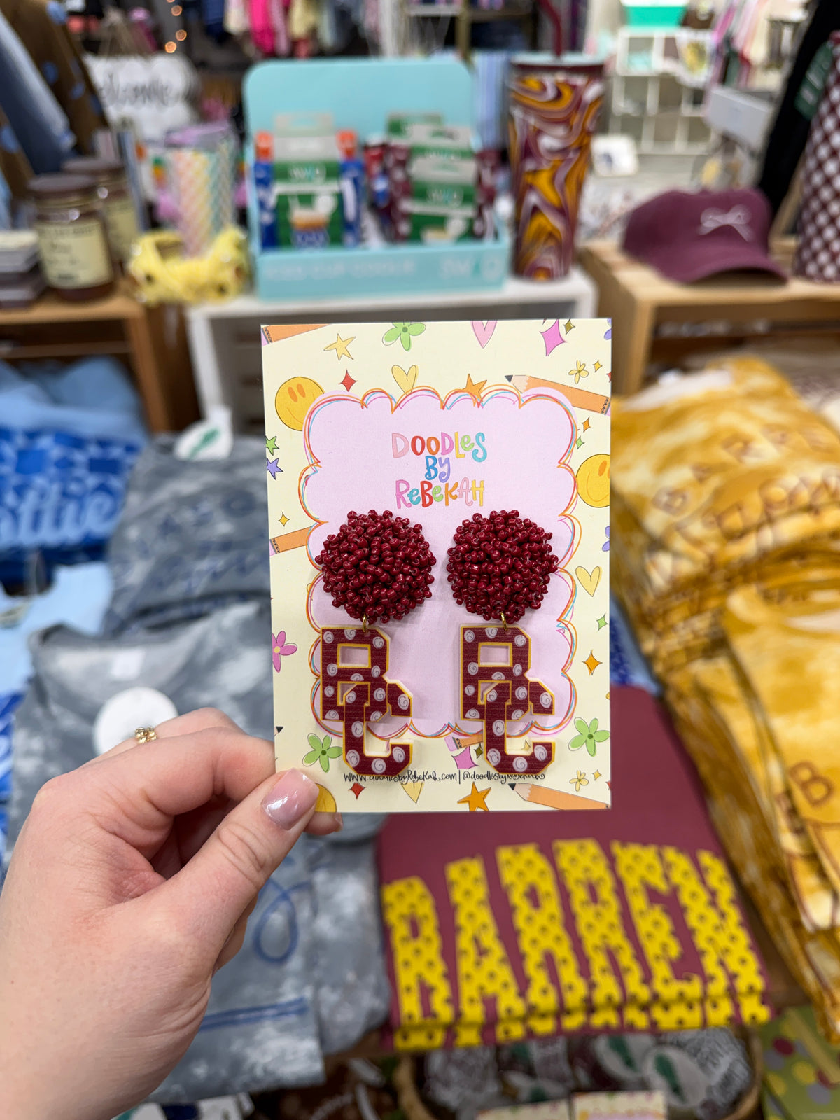 A hand holding a small display card featuring a pair of earrings: with deep red pom-pom studs and BC letters. The card reads 'Doodles By Rebekah”