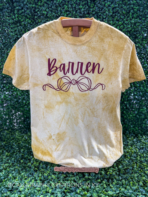 A short-sleeve crewneck t-shirt with a yellow and cream mineral-wash finish, featuring the name 'Barren' and a bow design in dark burgundy on the front, displayed on a wooden hanger against a green hedge background.