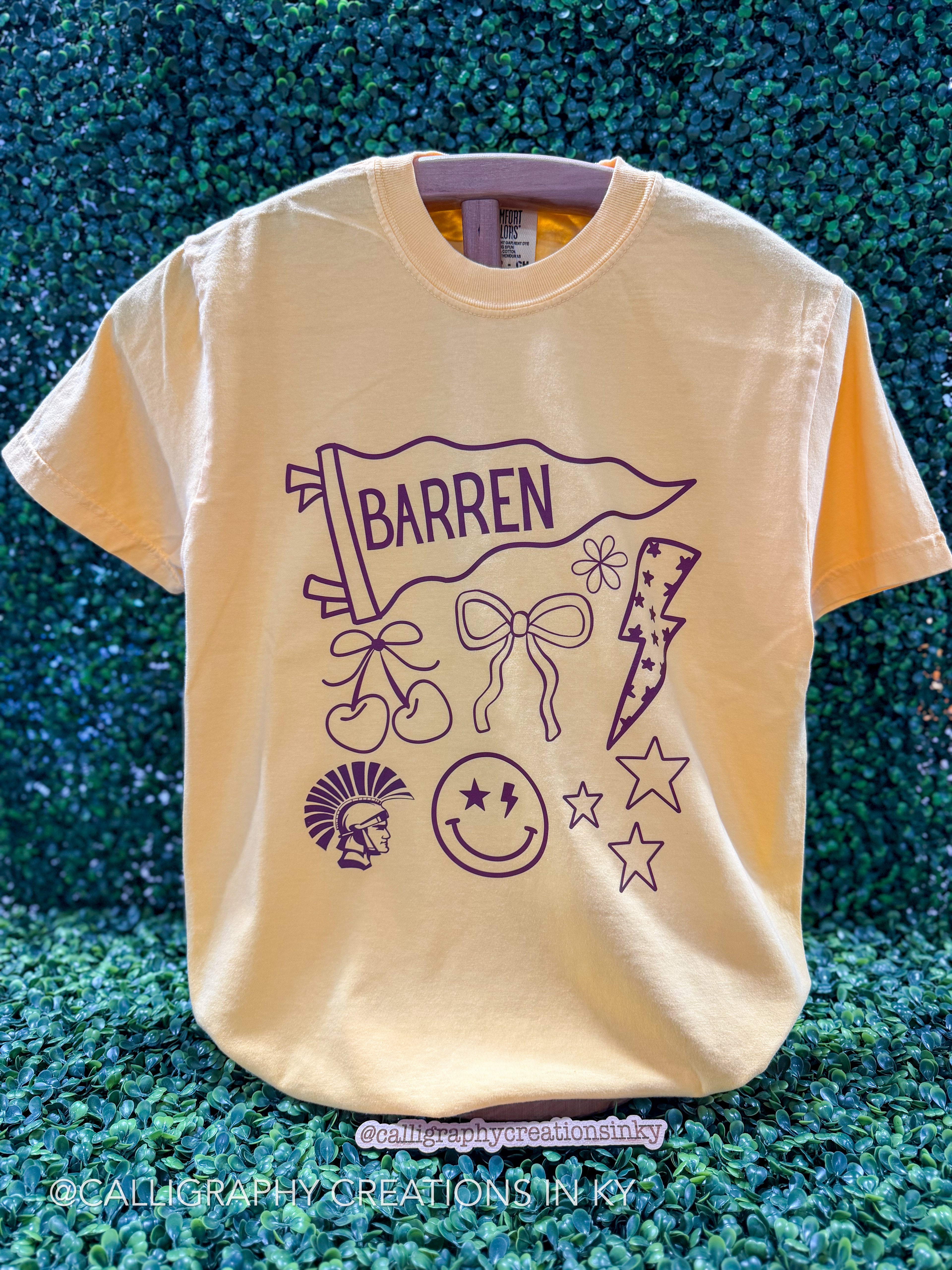 A pale yellow graphic t-shirt featuring a purple line art design with the word 'BARREN' on a pennant flag, bows, stars, a lightning bolt, and a smiley face, displayed against a green textured background.