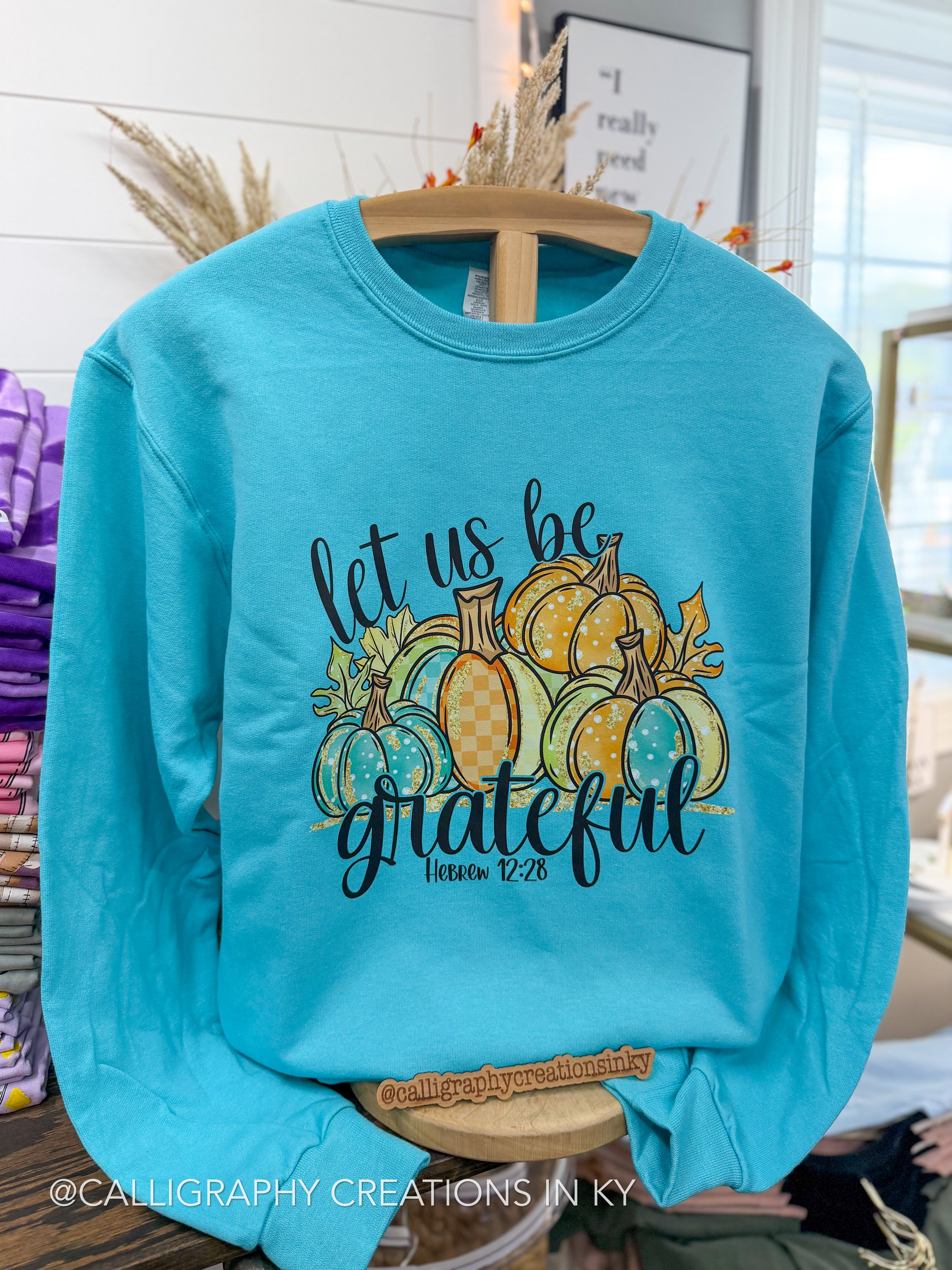 A turquoise crewneck sweatshirt hanging on a wooden hanger, featuring a fall-themed graphic with the text 'let us be grateful' and a colorful illustration of pumpkins. The sweatshirt is displayed in a retail setting.