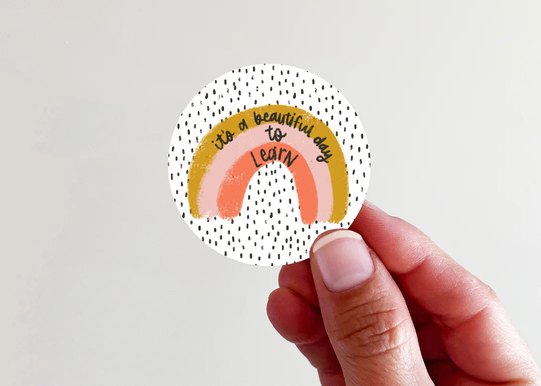 A hand holding a round sticker with a hand-drawn rainbow, black speckled background, and the text 'it's a beautiful day to LEARN'.