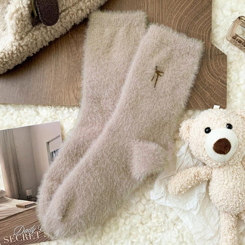 Beige socks with a brown embroidered bow near the cuff. With a teddy bear on a white fleece blanket in the background.