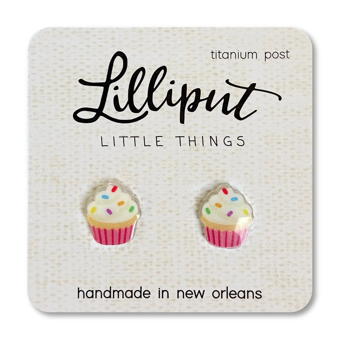 Cupcake earrings with pink liner, white frosting, and rainbow sprinkles on beige card on white background