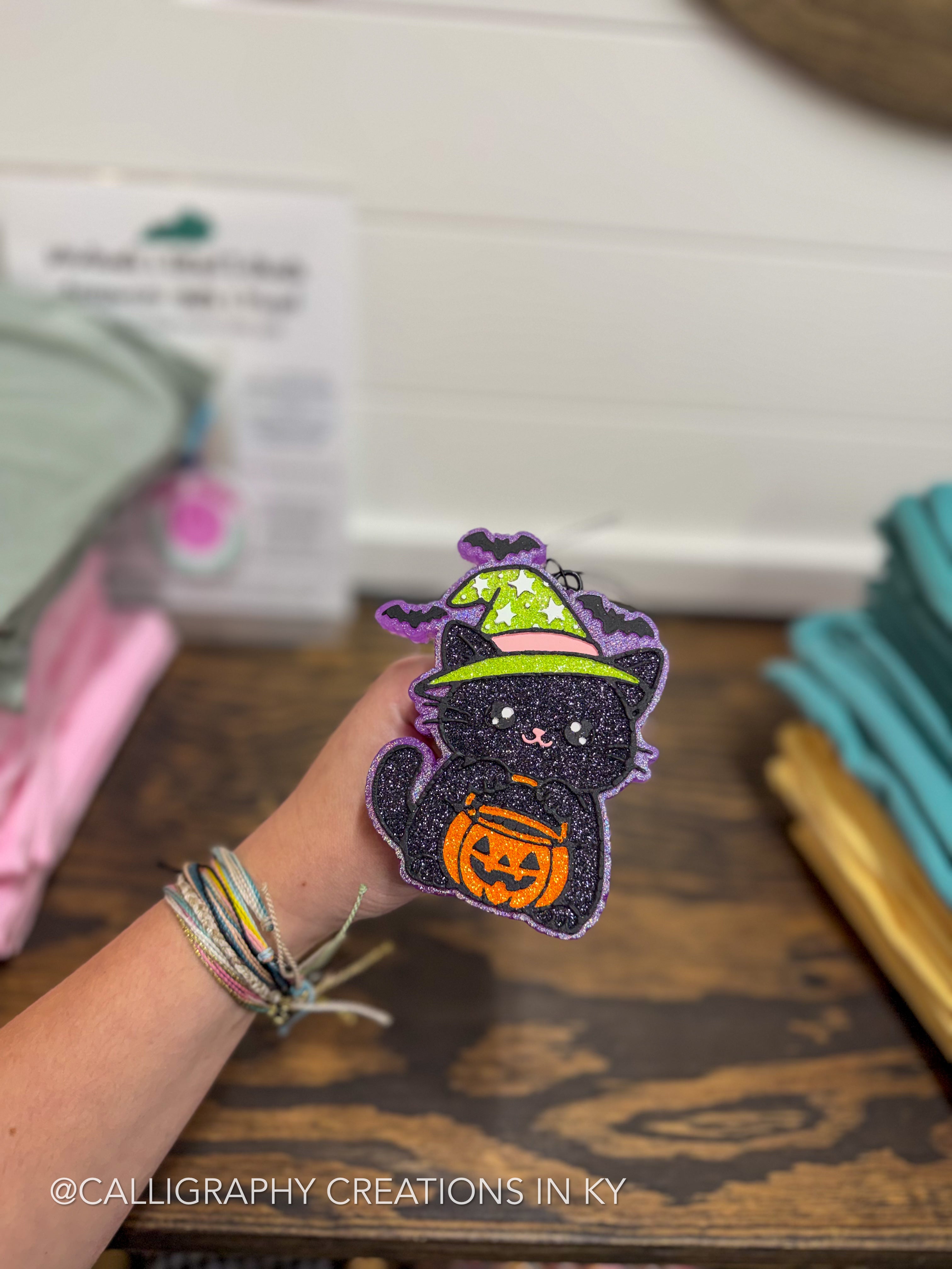 A hand holds a glittery black cat patch wearing a green witch hat and holding an orange pumpkin bucket, with a blurred craft shop background.