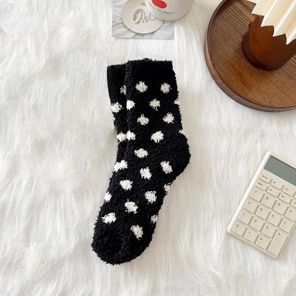 A single black fuzzy sock with white pom-pom dots, laid flat on a white faux fur surface with styled props including a wooden tray, a calculator, and a cup.