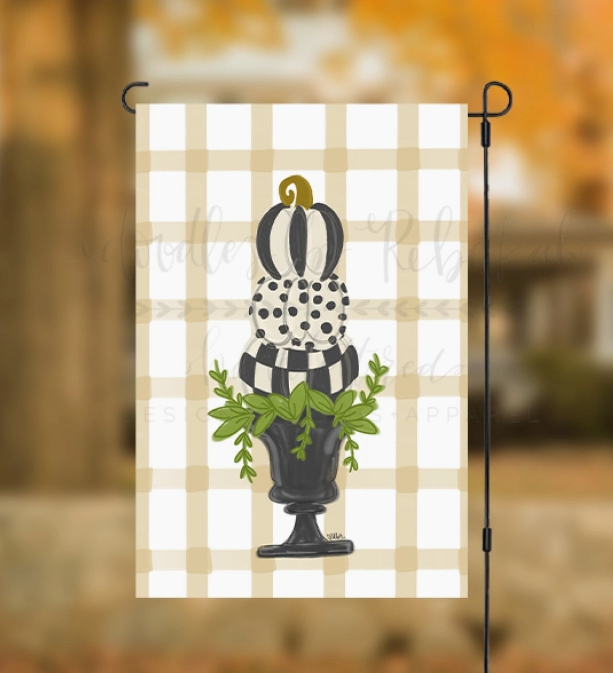A vertical garden flag hanging from a black metal stand, featuring a black and white gingham background with a central illustration of a black and white pumpkin, a polka-dot pumpkin, a striped pumpkin, and green leaves in a black pot. The background is a blurred outdoor scene with warm autumn tones.