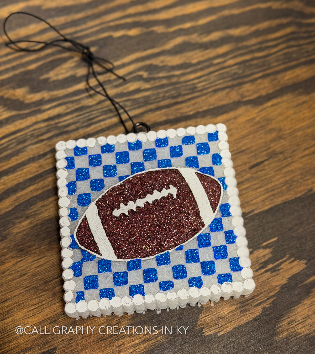 A square freshie featuring a blue and white checkered pattern with a glittery brown football in the center, hanging on a wooden surface.