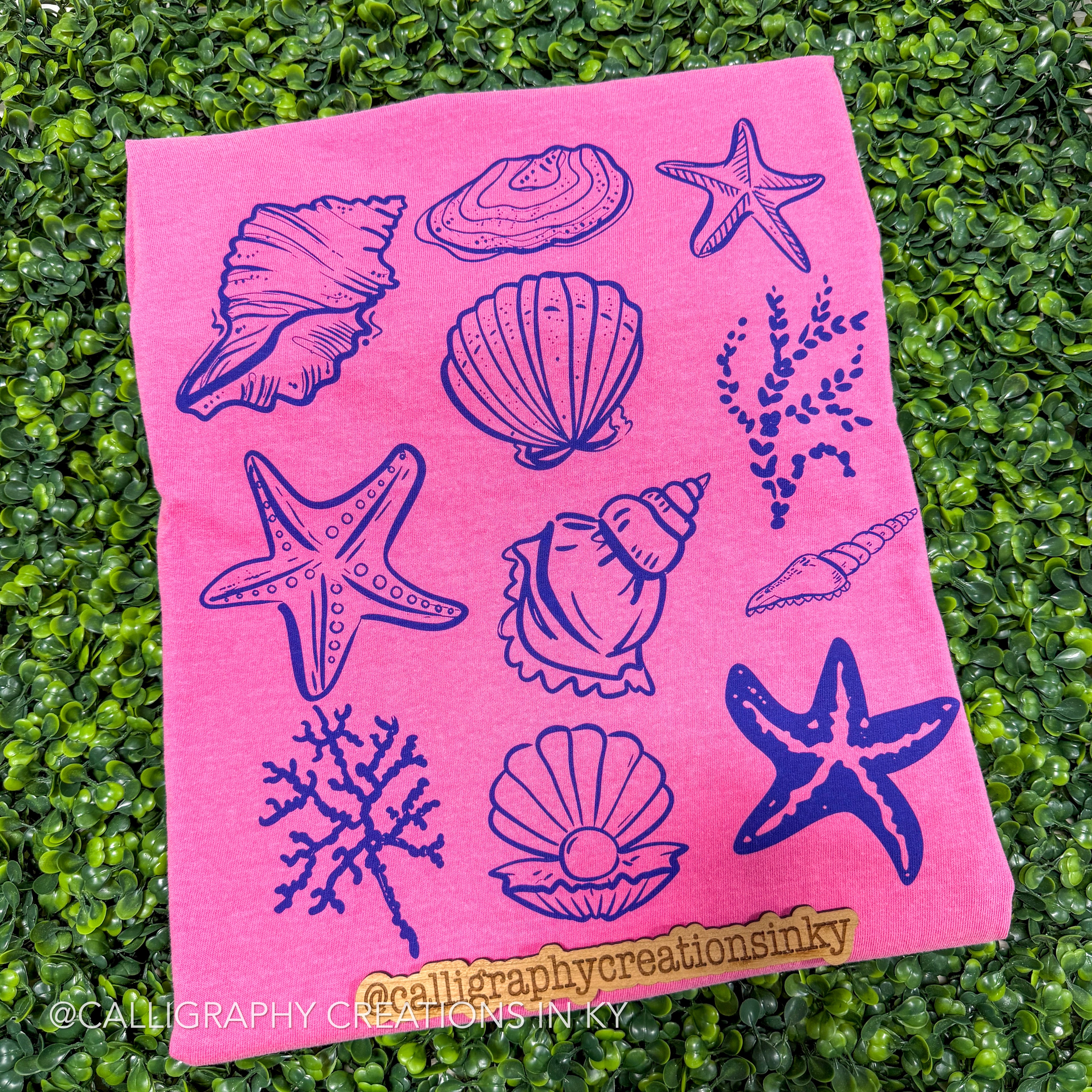 Pink t-shirt with blue sea-themed designs on a green grass background