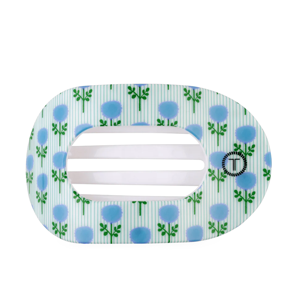 A hair claw clip with a white and light blue striped background, decorated with green leafy stems and blue dandelion-like flowers. The center has three horizontal white bars, and a small circular brand logo with a black 'T' is visible on the right side.