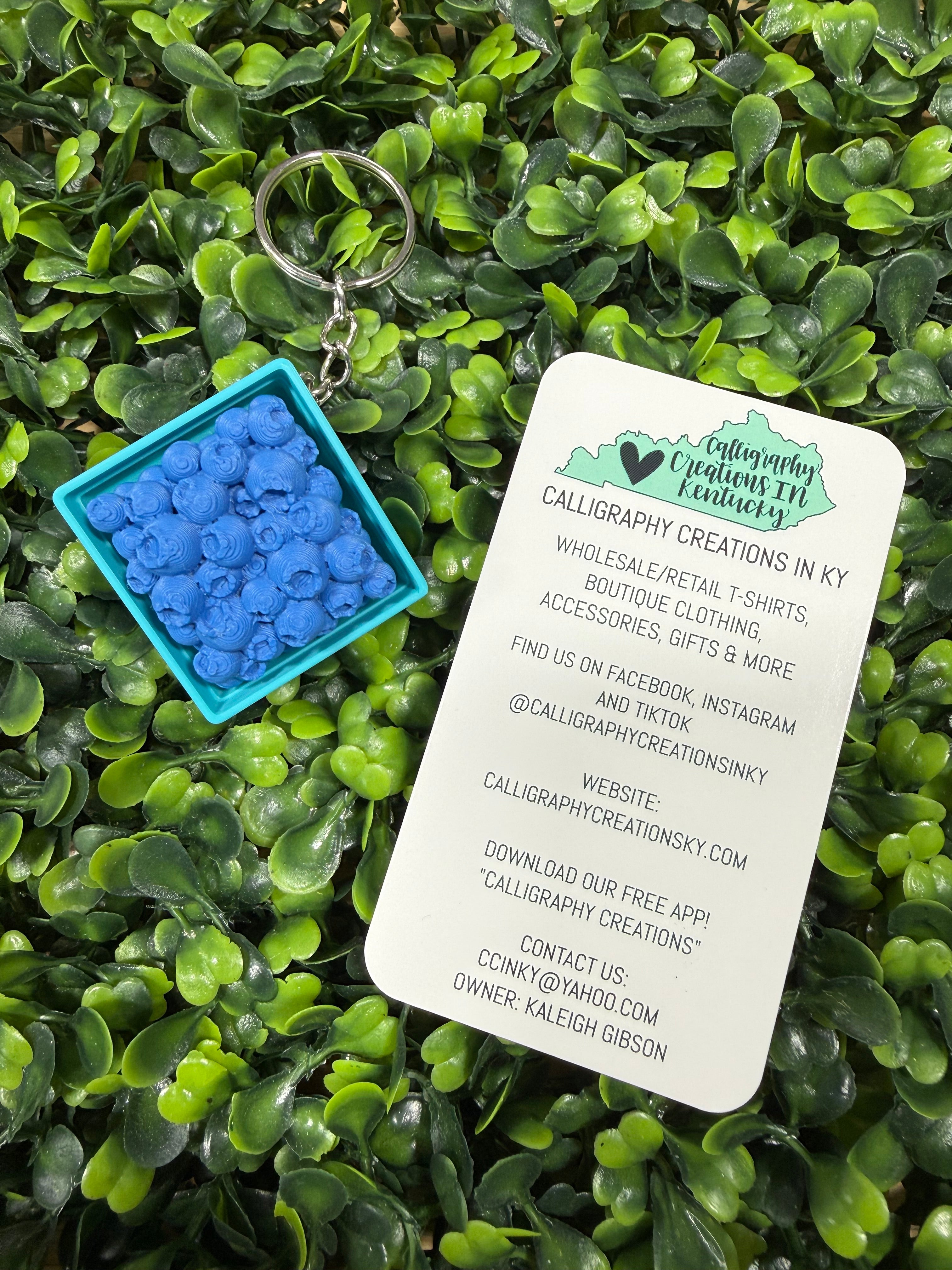 A small square turquoise container filled with bright blue flower-shaped calligraphy charms, placed on green foliage next to a white printed card with branding and contact details.