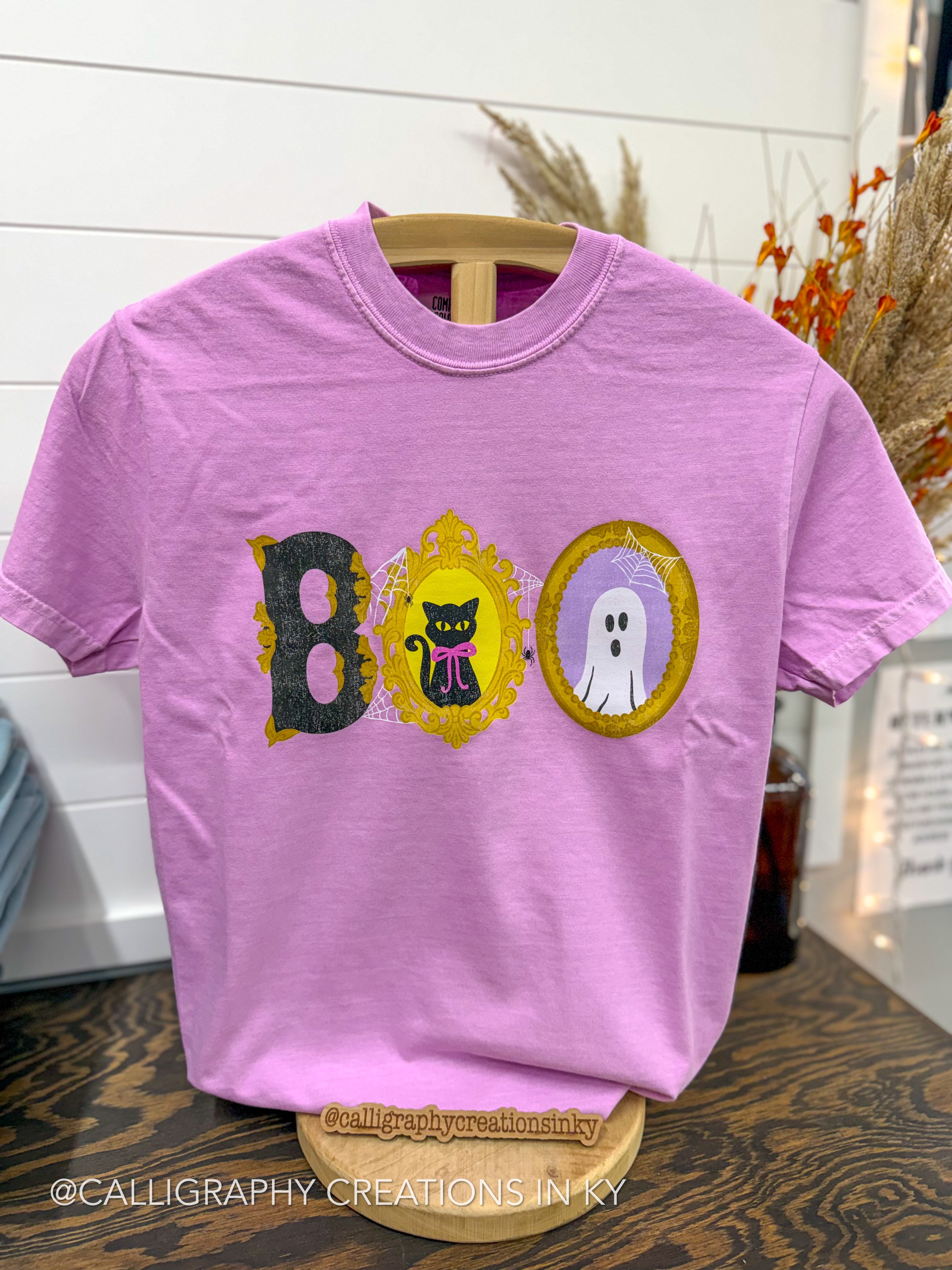A light purple short-sleeve t-shirt displayed on a wooden hanger stand. The front features a 'BOO' graphic spelling out Halloween-themed shapes: a black cat in a gold frame for the 'B', a black cat with a pink bow in a gold frame for the 'O', and a white ghost in a gold frame for the 'O'.