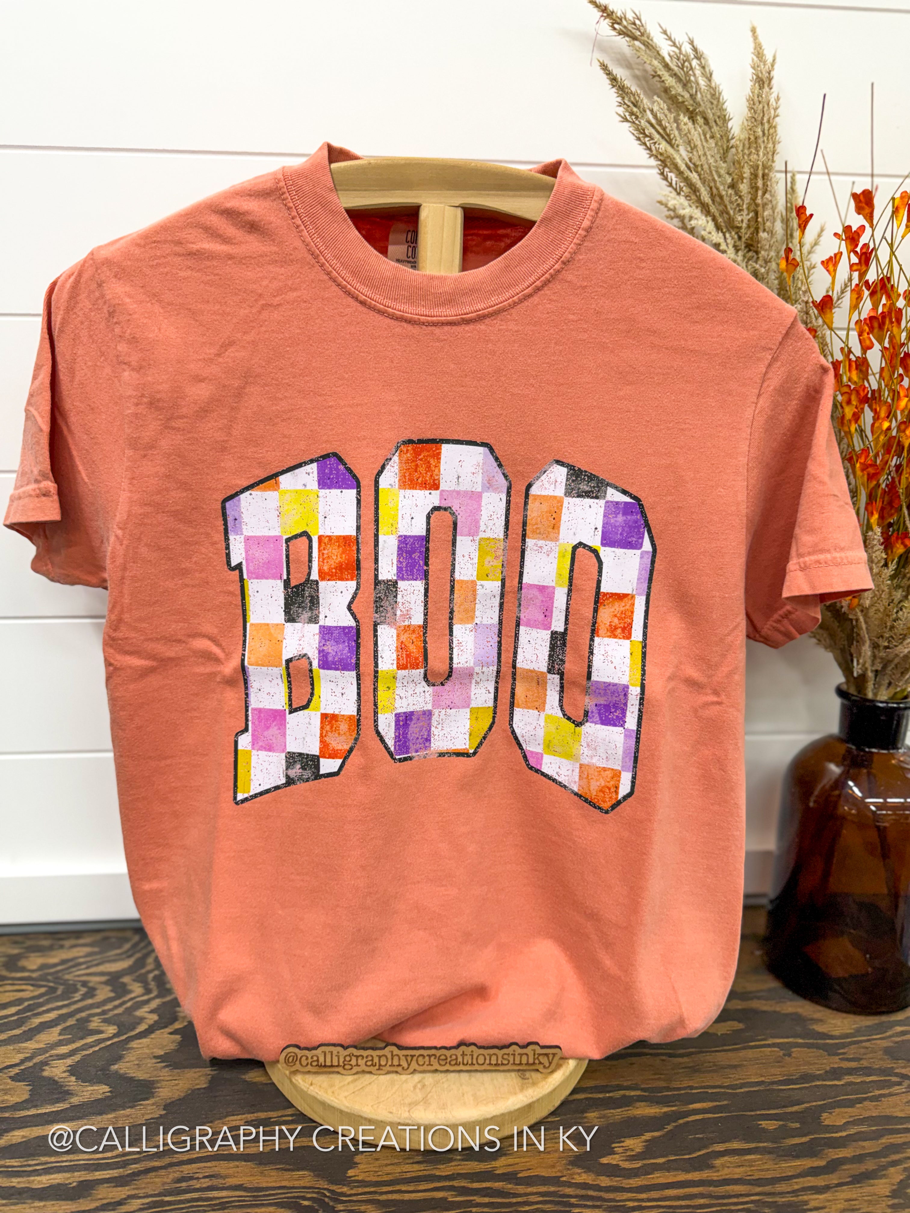 A coral orange crewneck t-shirt displayed on a wooden hanger. The front of the shirt features a large graphic spelling 'BOO' in a retro collegiate font, filled with a multicolor patchwork checkerboard pattern of orange, purple, yellow, white, and black.