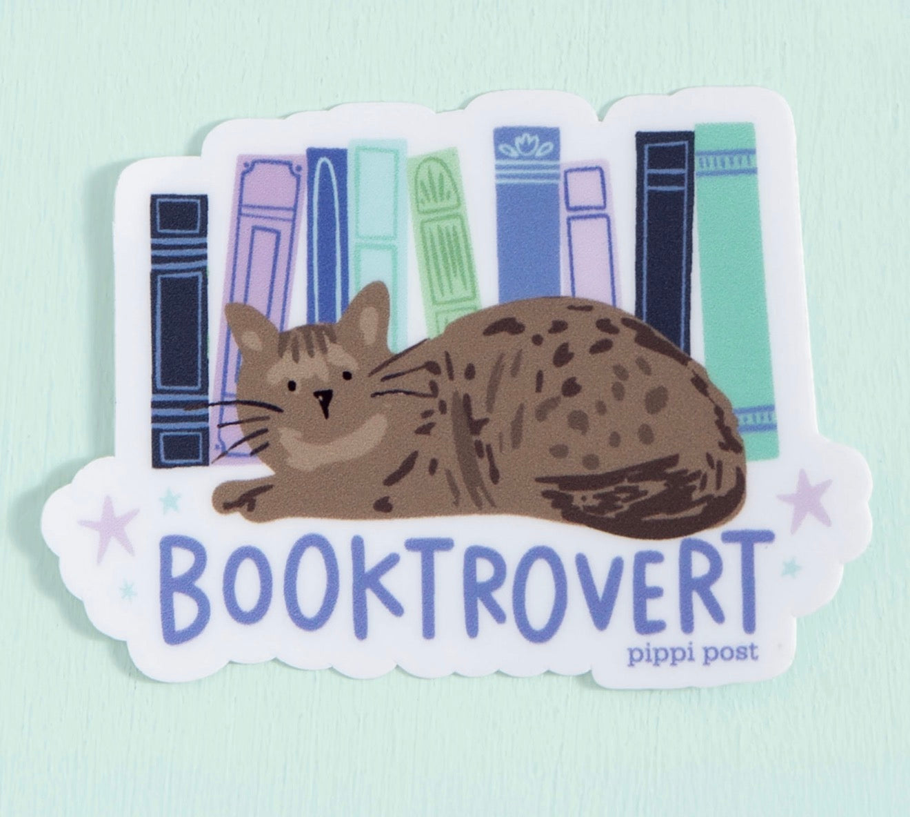 A die-cut sticker featuring a brown tabby cat curled up in front of illustrated book spines, with the word 'BOOKTROVERT' in blue-purple text and 'pippi post' printed below, shown on a pale mint background.