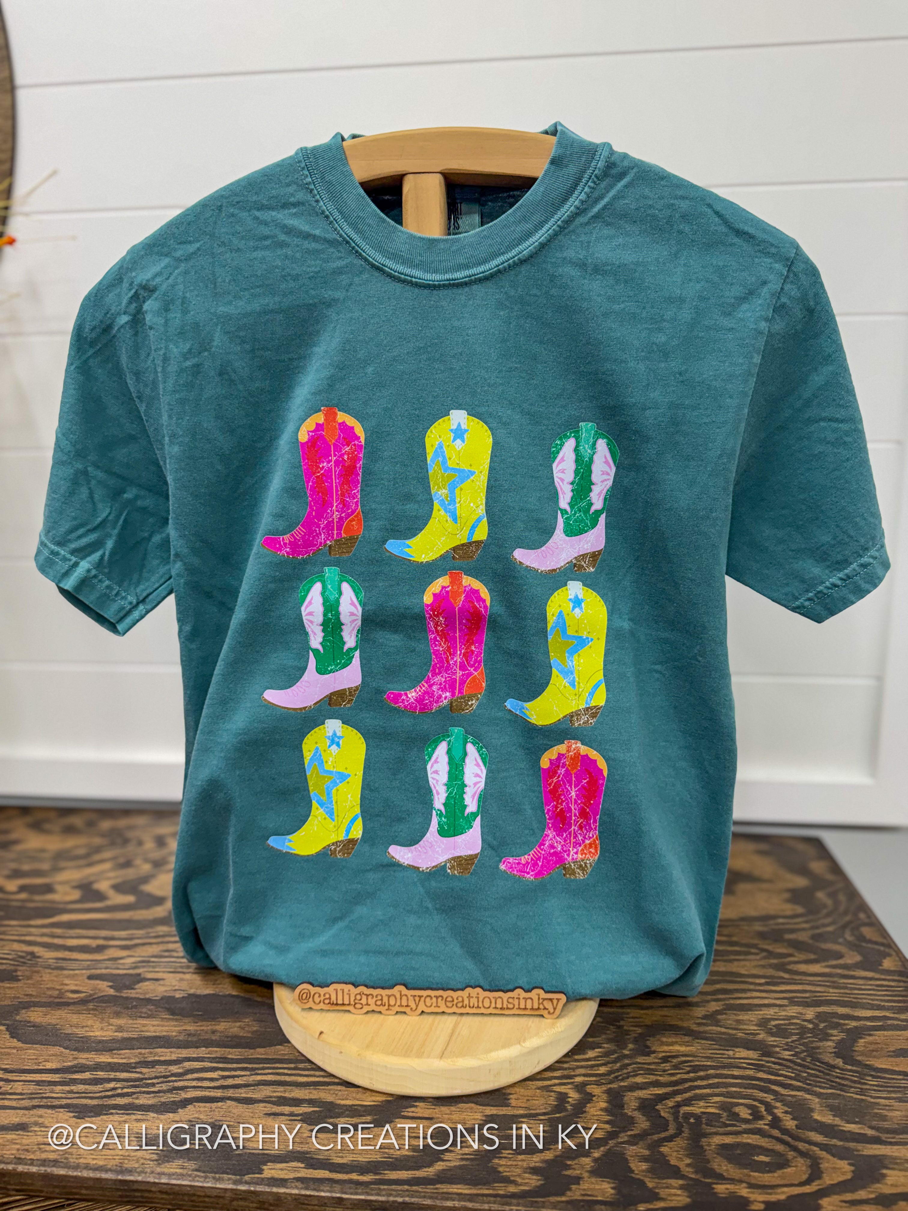 A teal short-sleeve t-shirt displayed on a wooden hanger stand, featuring a graphic print of nine colorful cowboy boots in a grid pattern. The background consists of a white paneled wall and a dark wood table.