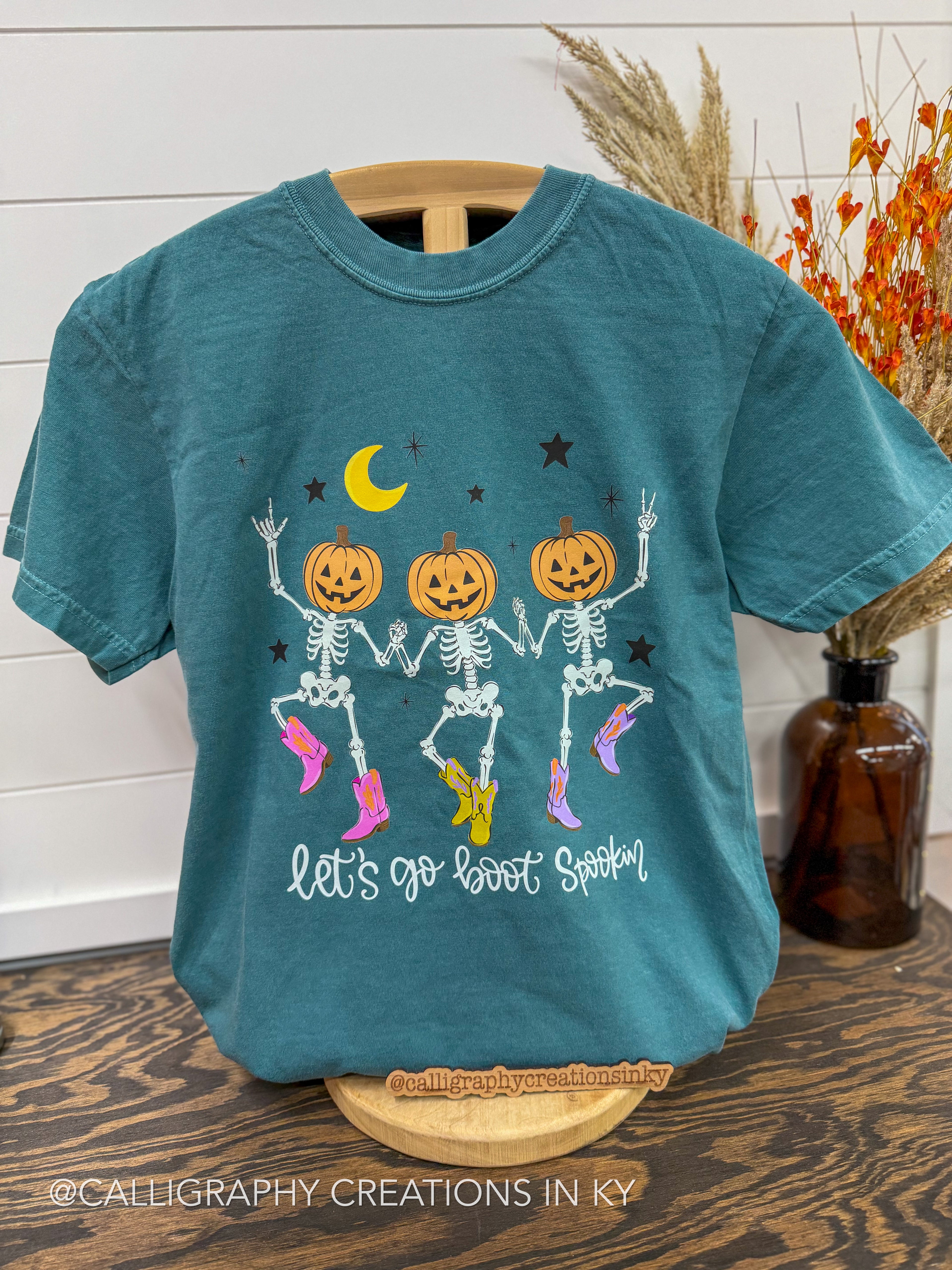 A teal blue short-sleeve t-shirt displayed on a wooden hanger stand, featuring a festive graphic of three dancing skeleton pumpkins under a crescent moon and stars, with the text 'let's go boot spookin'' below. The shirt is styled on a wooden table next to a brown glass bottle and dried grass.