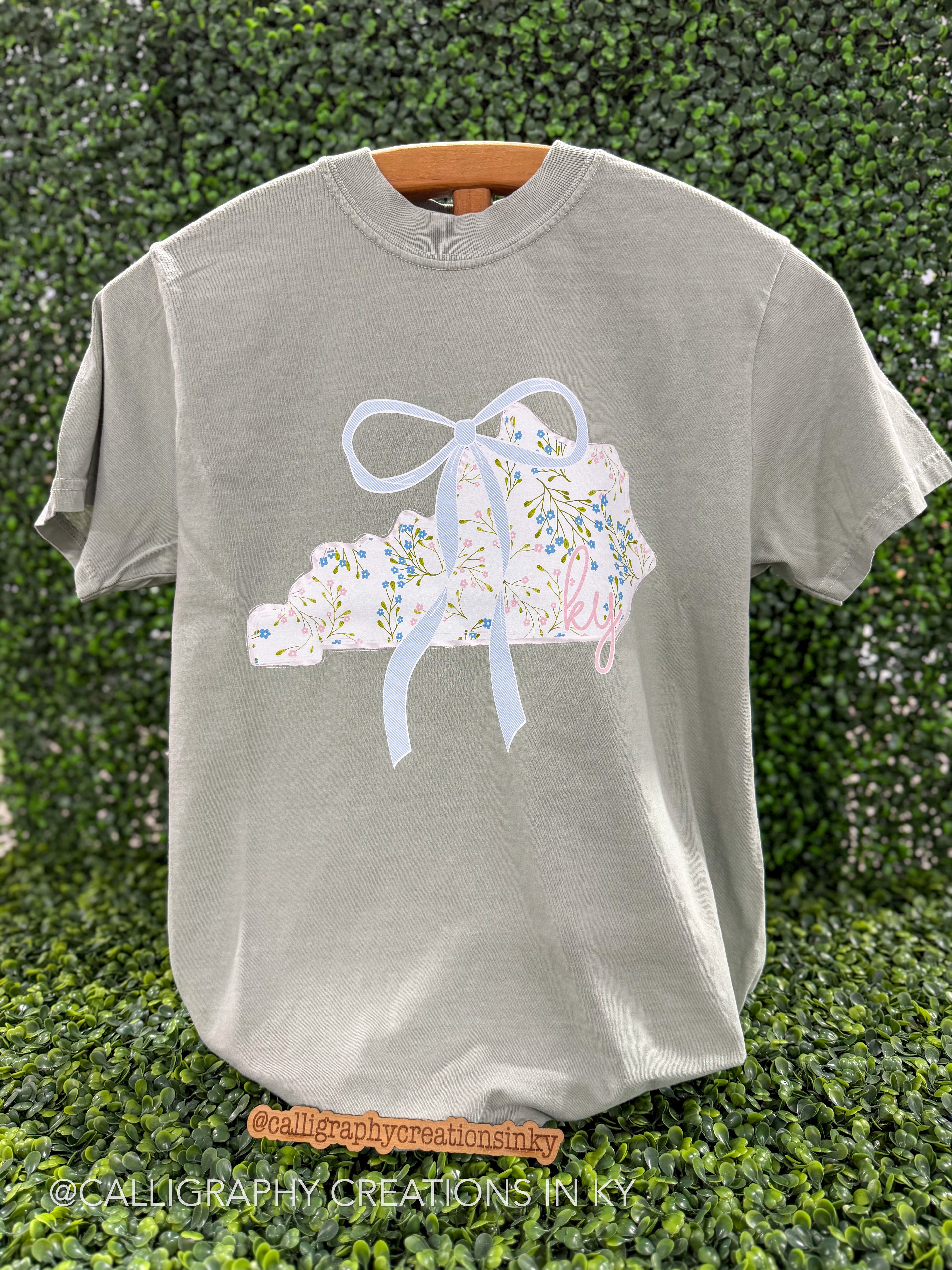Sage green shirt with floral Kentucky shaped pattern on a wooden stand