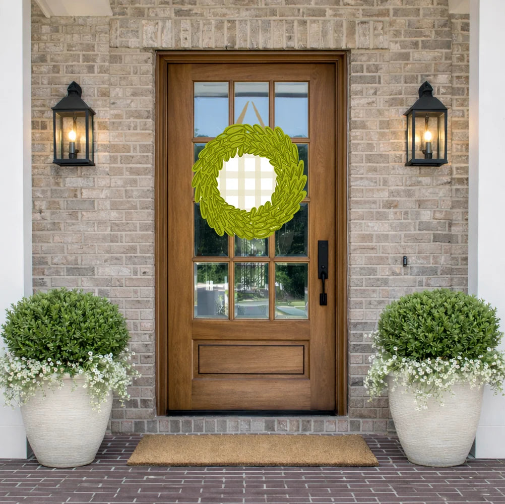 A green woven wreath with a white cross in the center hangs on a brown wooden front door, flanked by two black wall lanterns and potted green shrubs on a brick porch.
