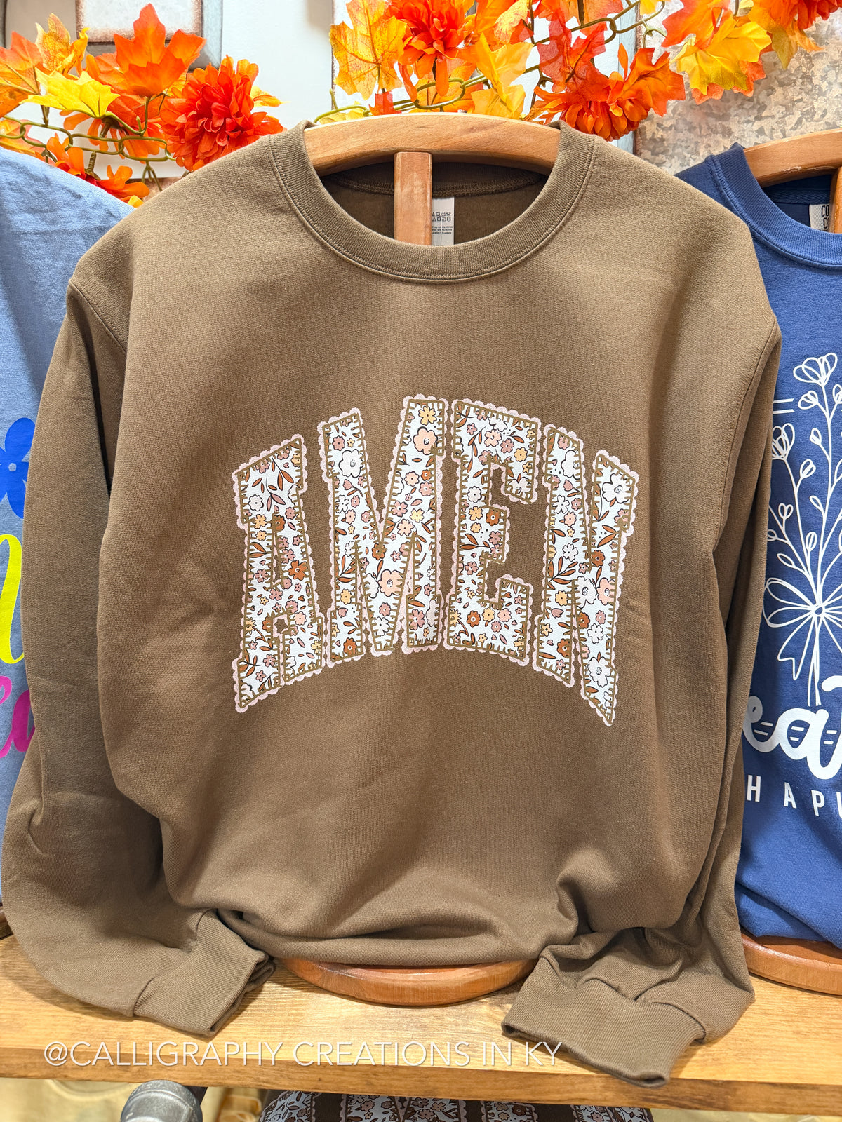 A brown crewneck sweatshirt featuring the word 'AMEN' in an arched collegiate font, filled with a white floral pattern. The sweatshirt is displayed on a wooden hanger on a wooden shelf, with other colorful shirts and autumn leaves in the background.