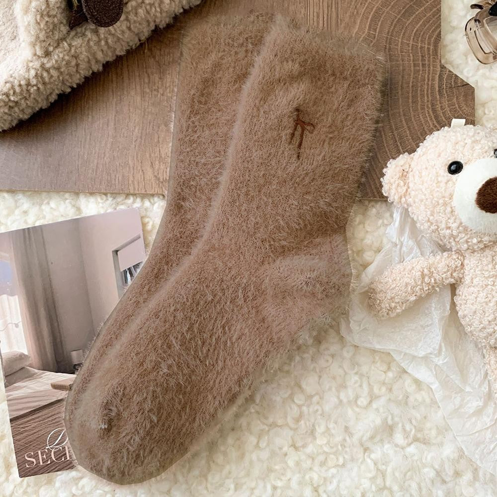 A pair of plush, fuzzy ankle socks in a warm taupe-brown color, styled in a cozy flat-lay arrangement with a teddy bear, a wooden slice, and a small inset photo.