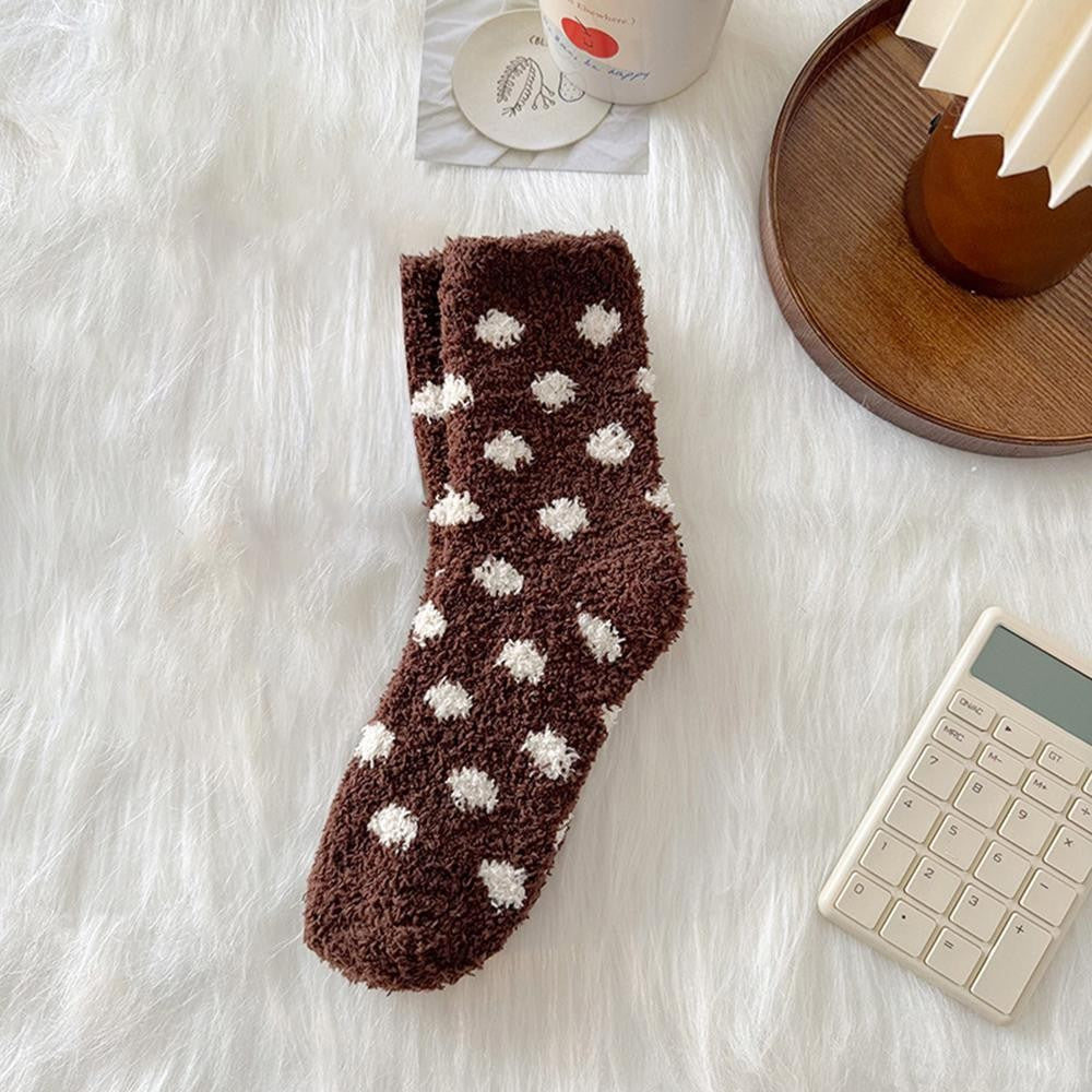 A single brown fuzzy sock with white polka dots is laid flat on a white faux-fur surface, surrounded by styled props including a wooden tray, a calculator, and a cup.