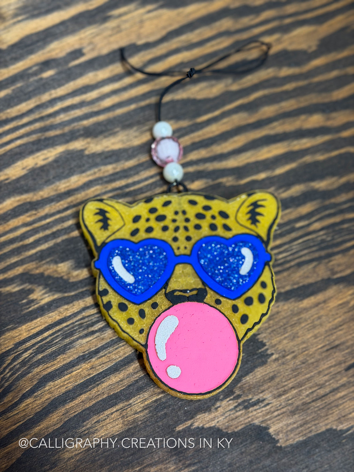 A handcrafted freshie car charm shaped like a leopard head with yellow and black spotted fur, blue heart-shaped sunglasses, and a large pink bubblegum bubble, resting on a dark wood background.