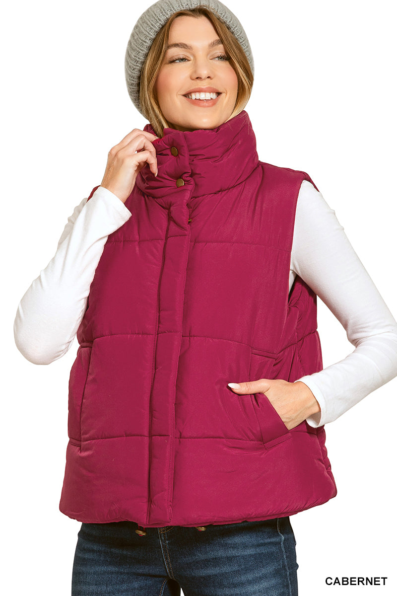 A woman wearing a sleeveless quilted puffer vest in a deep cabernet burgundy color, styled over a white long-sleeve shirt and blue jeans, with a gray knit beanie. The word 'CABERNET' is printed in the bottom right corner.