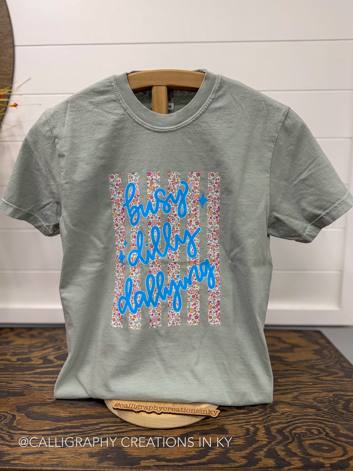 A sage green crewneck t-shirt on a wooden hanger, featuring a graphic design with blue cursive text reading 'buzzy dippy dallying' over vertical stripes of pink, white, and blue floral patterns. The shirt is displayed on a dark wood table against a white shiplap wall.