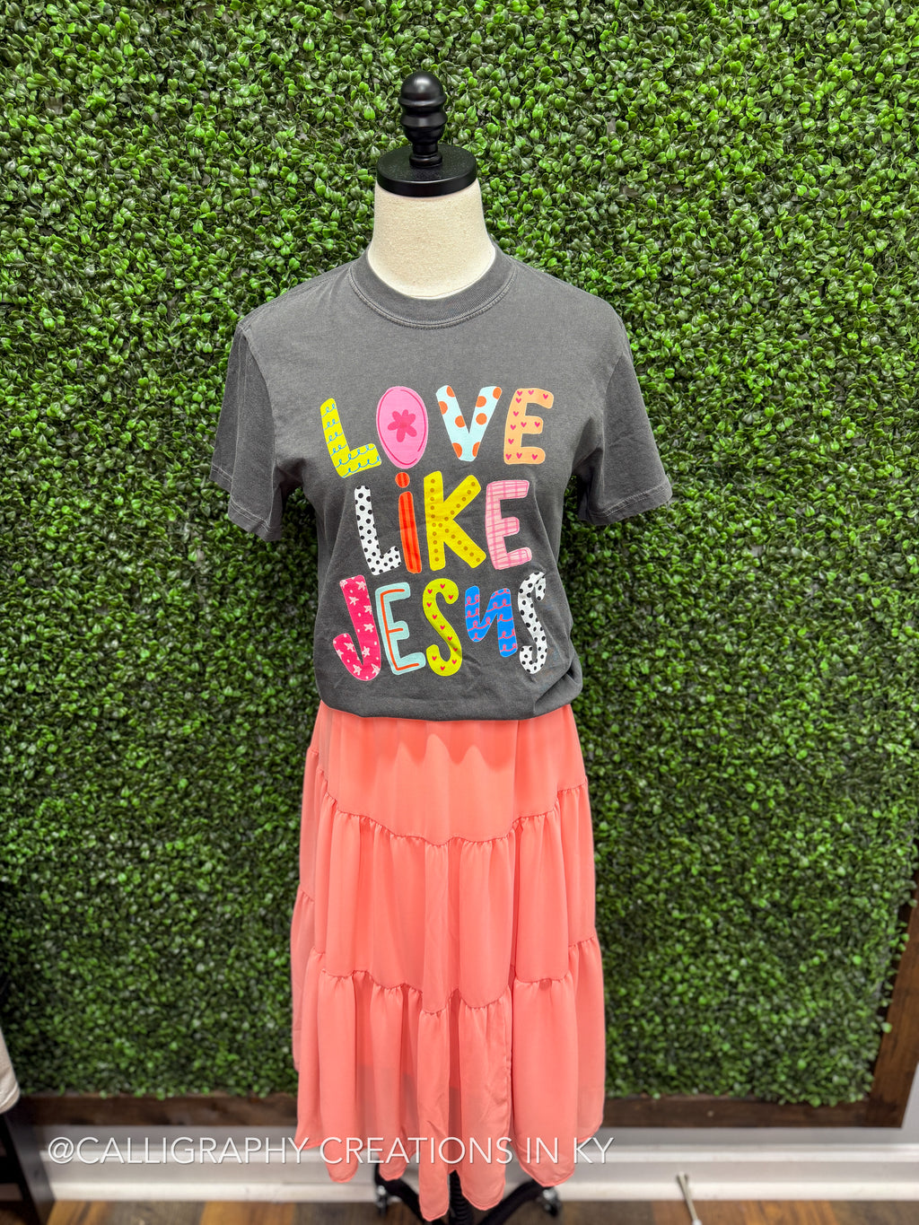 Mannequin wearing a gray t-shirt with colorful text and a peach skirt against a green leafy background.