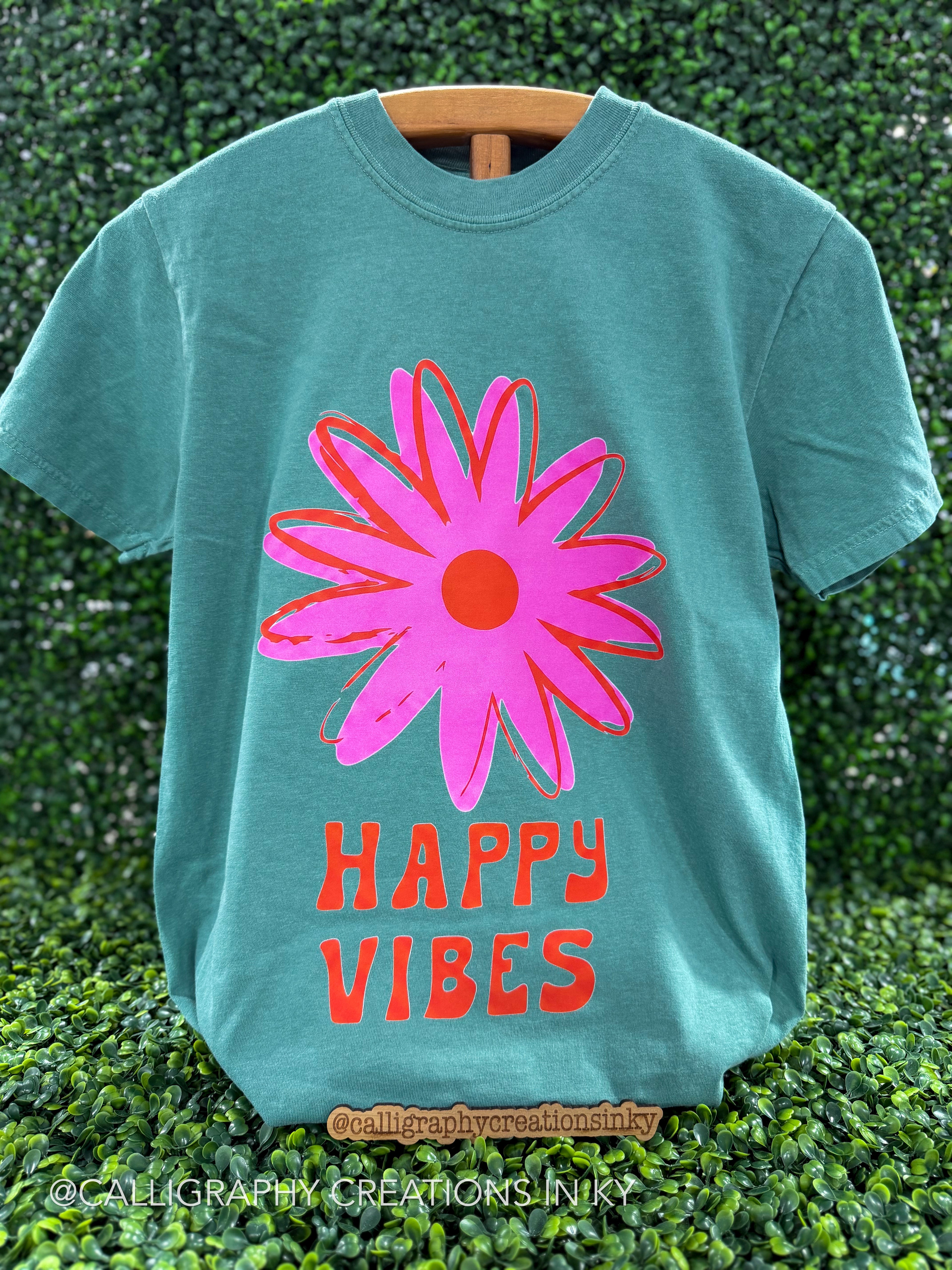 Light green tee with a flower design that says “happy vibes”