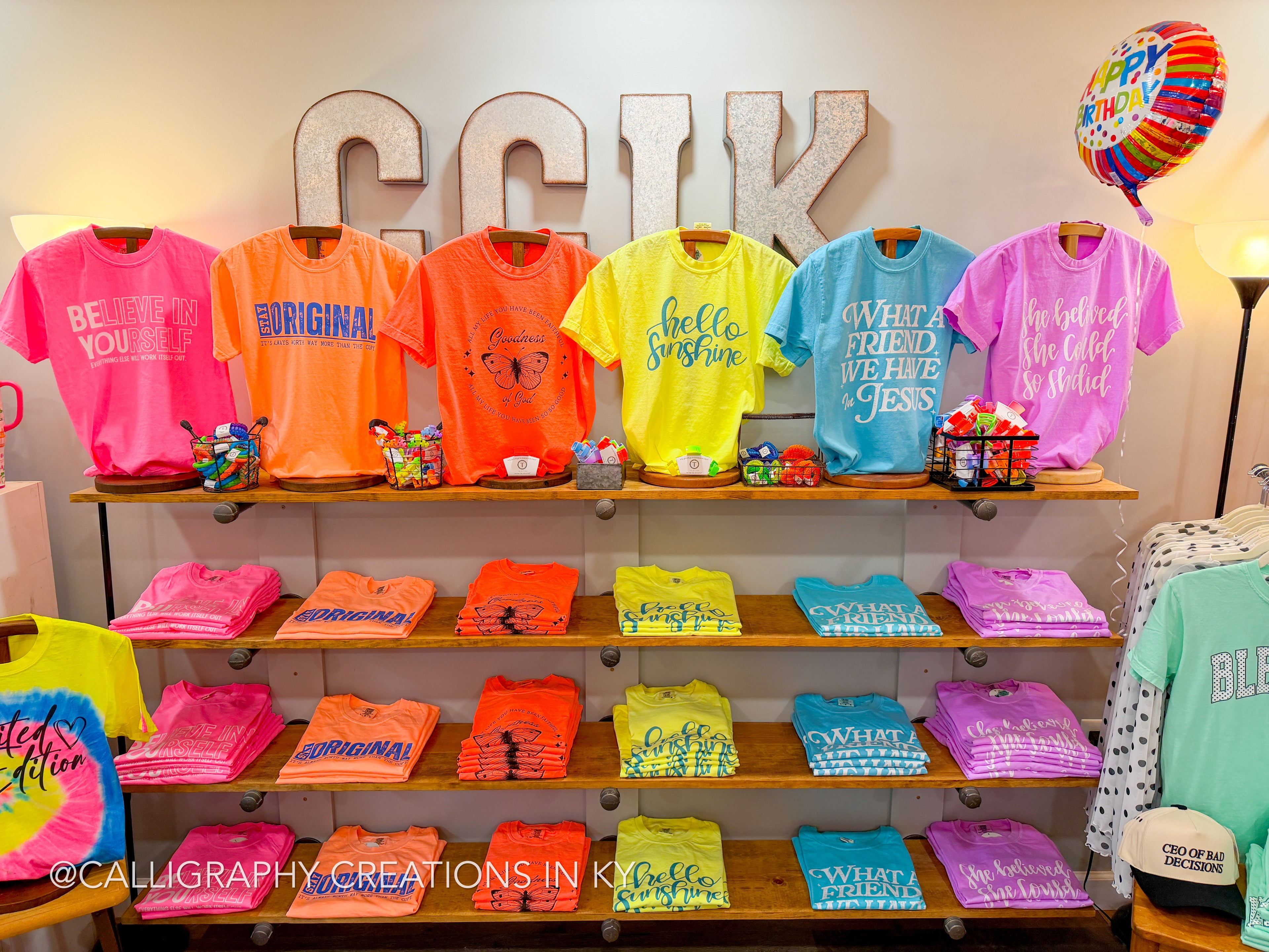 Display of colorful t-shirts on shelves with Calligraphy Creations in Kentucky branding.