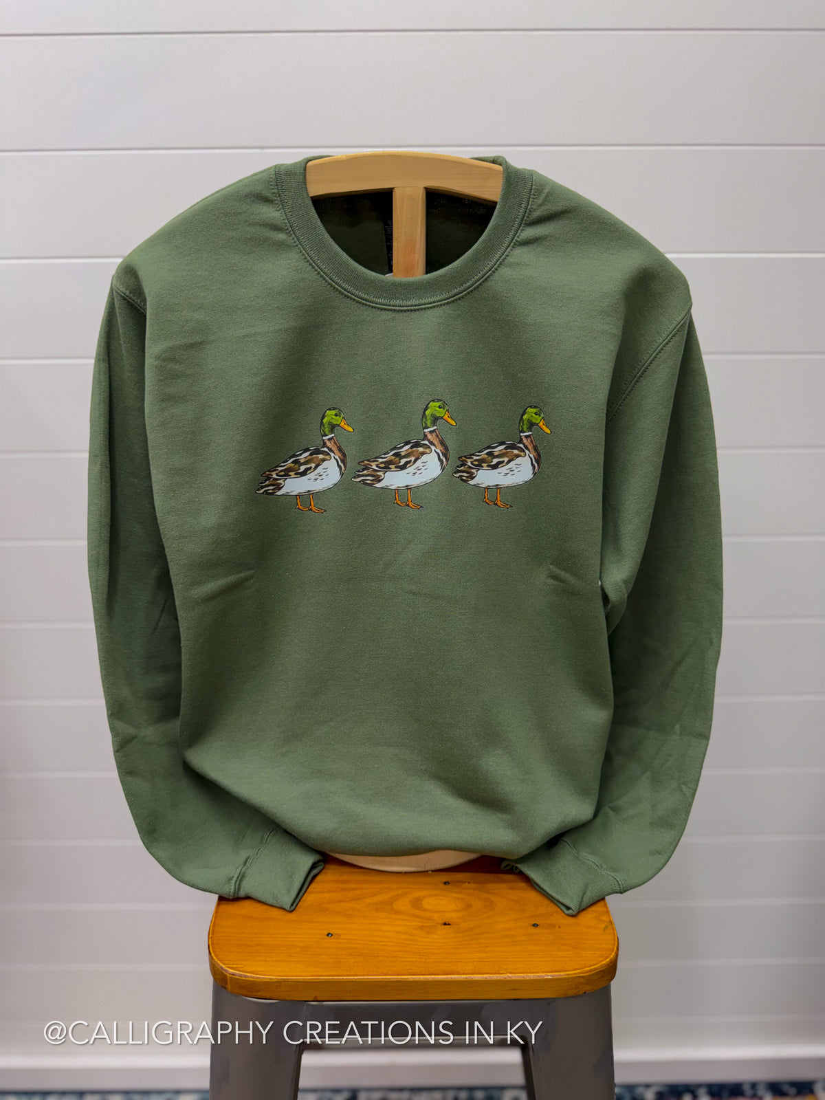 An olive green crewneck sweatshirt featuring an embroidered row of three mallards across the chest, displayed on a wooden hanger draped over a wooden stool against a white shiplap wall.