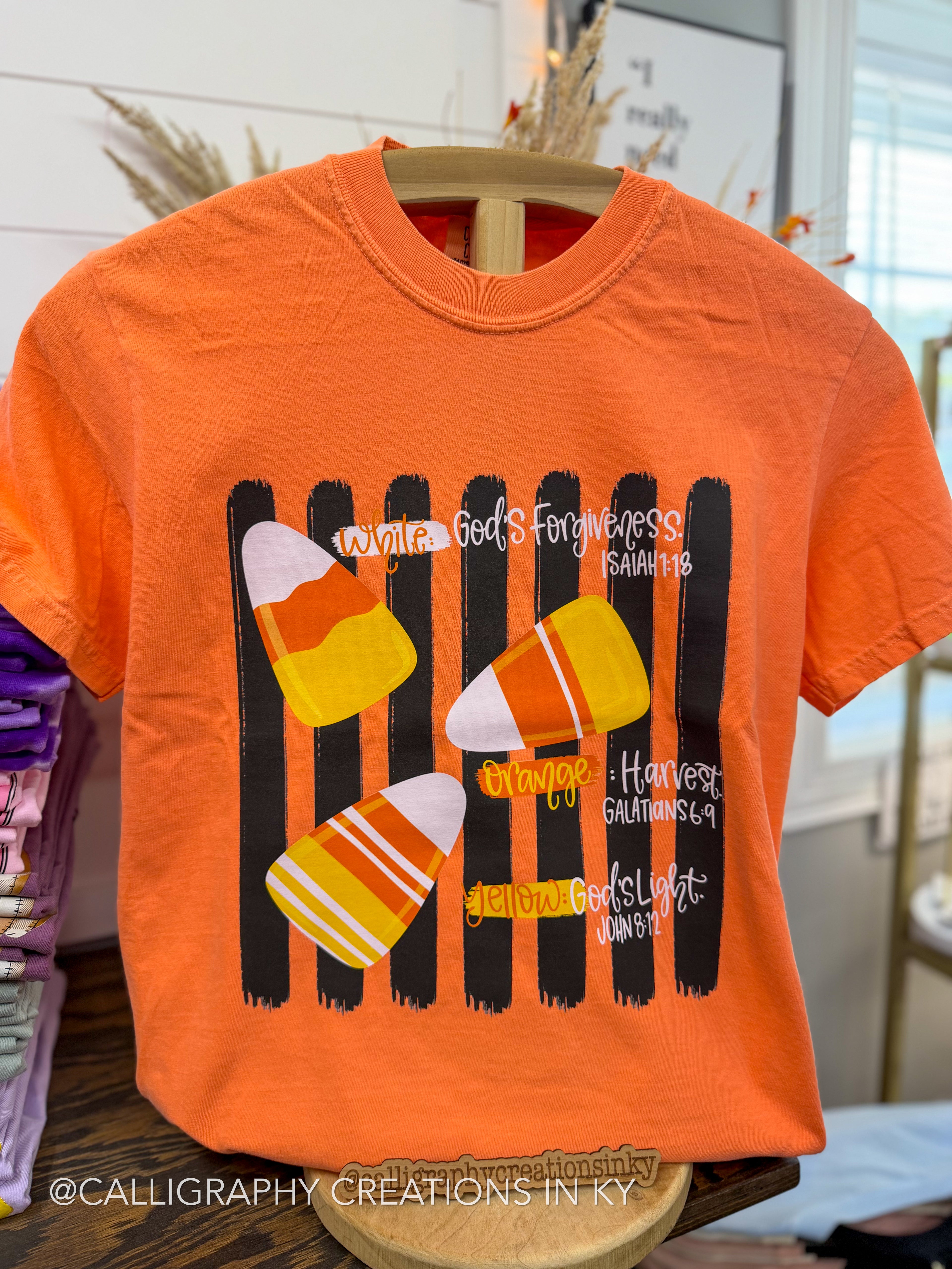 An orange short-sleeve graphic T-shirt displayed on a wooden hanger. The shirt features a Halloween-themed design with four candy corn illustrations over black vertical stripes, surrounded by white script text reading 'Harvest', 'Forgiveness', 'Orange', 'Yellow', and 'Light'.